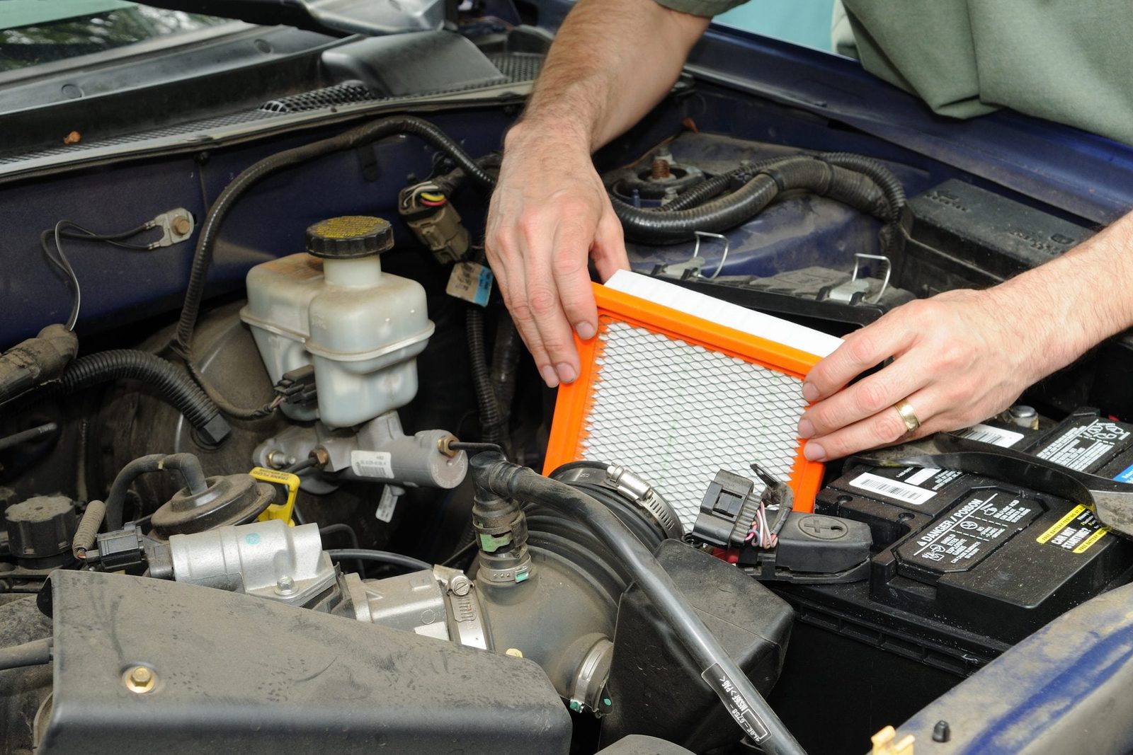 A person installs an orange-framed air filter in a vehicle's engine compartment, surrounded by various automotive components and a car batte