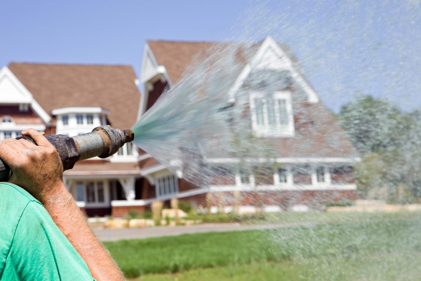 A person uses a garden hose to spray water, nurturing a yard in front of a large, well-maintained house under a clear blue sky.