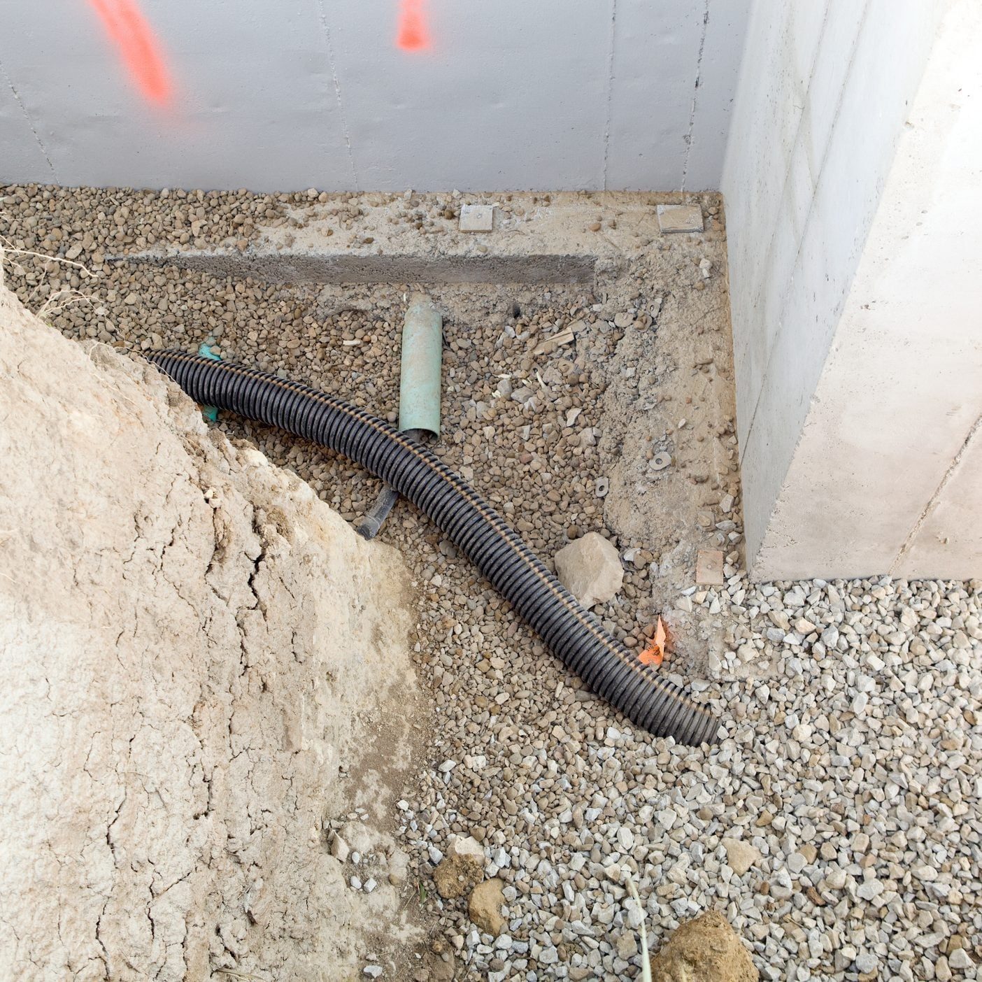 A flexible black pipe lies on gravel near a concrete foundation, surrounded by loose rocks and soil, with a green pipe and debris nearby.