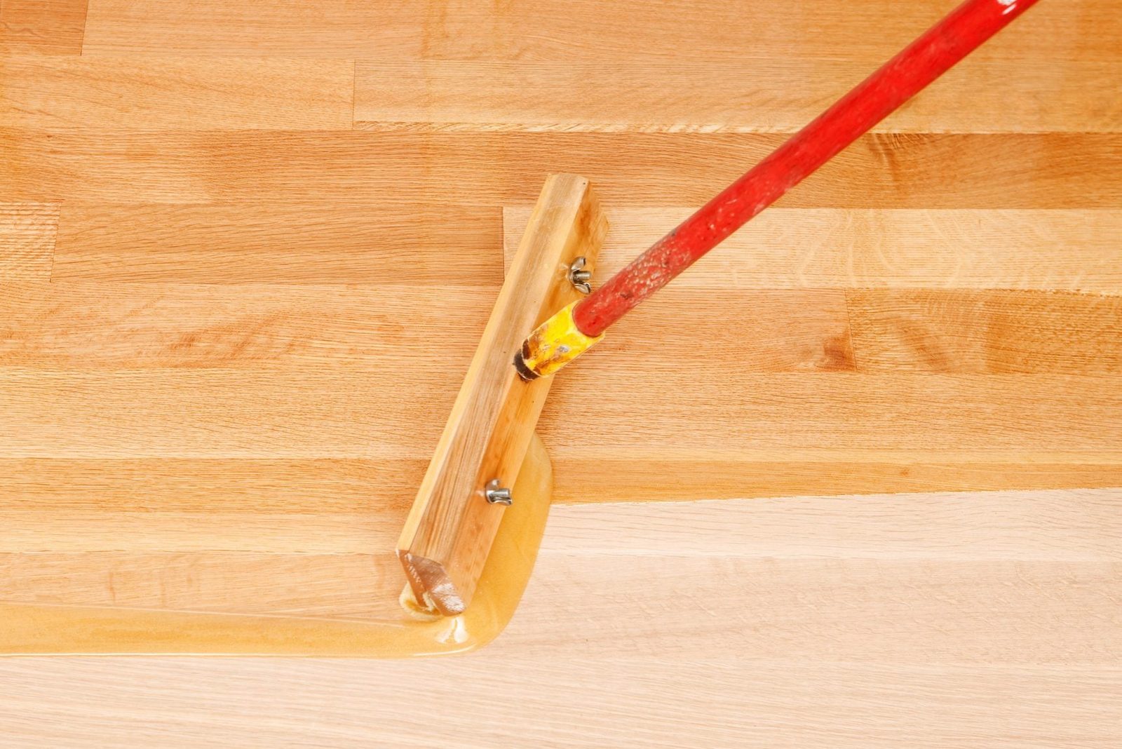 A wooden trowel spreads a yellow adhesive across a flat, polished wooden surface, demonstrating a smooth application in a workspace setting.