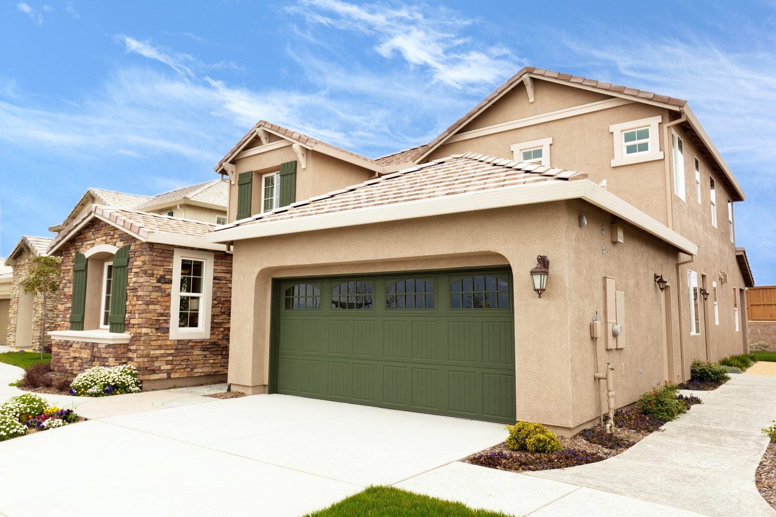 A suburban house features a stone facade, green garage door, and flower beds. It's set against a clear blue sky and well-maintained lawn.