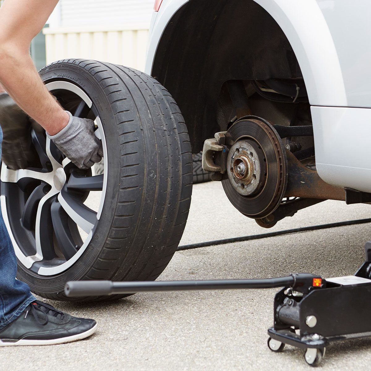 A person is replacing a tire on a car, using a jack on a concrete surface, with tools and the removed tire nearby.