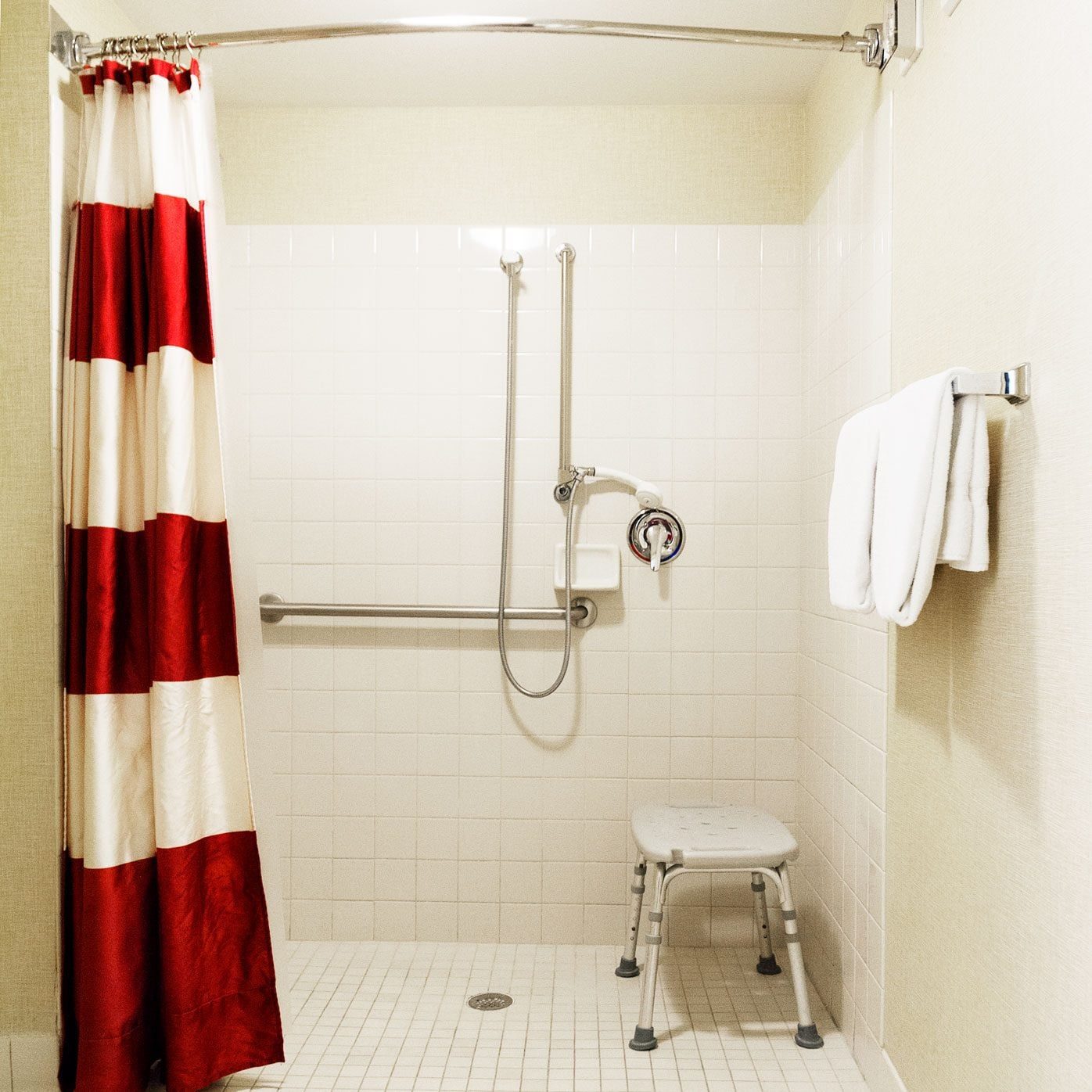A shower stall features a red-and-white striped curtain, a showerhead, grab bars, a stool, and neatly folded towels. Bright, tiled walls cre