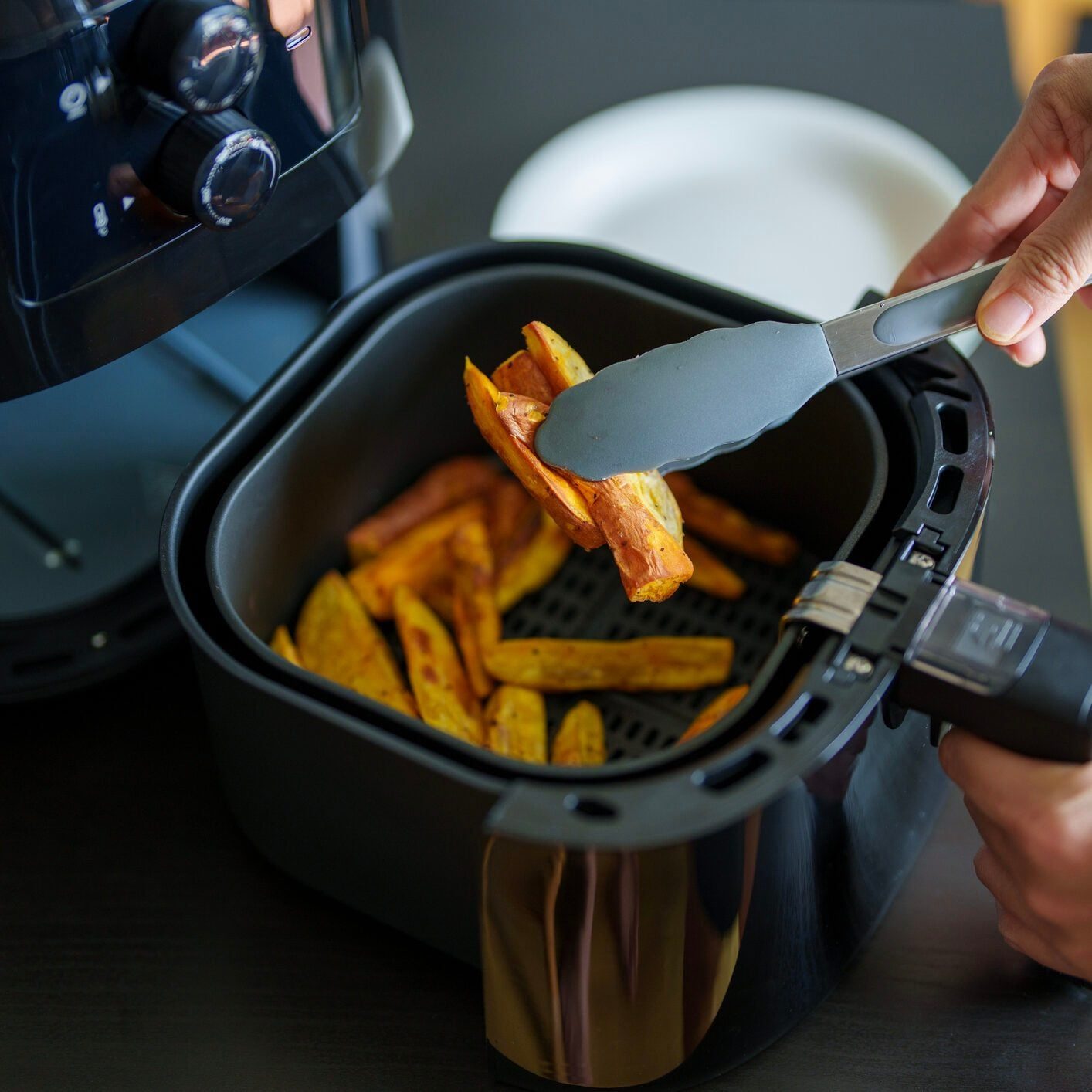 Tongs pick up pieces of potato wedges from the basket of an air fryer