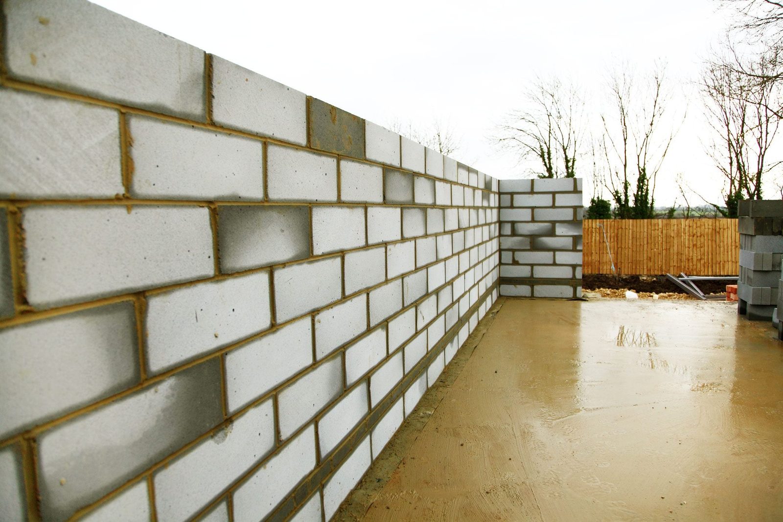 Gray concrete blocks form a partially constructed wall on a smooth floor, surrounded by trees and a wooden fence in a construction area.