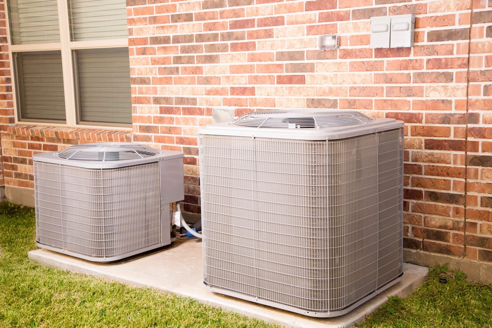 Two air conditioning units sit on a concrete slab beside a brick wall, surrounded by grass in a residential outdoor area.