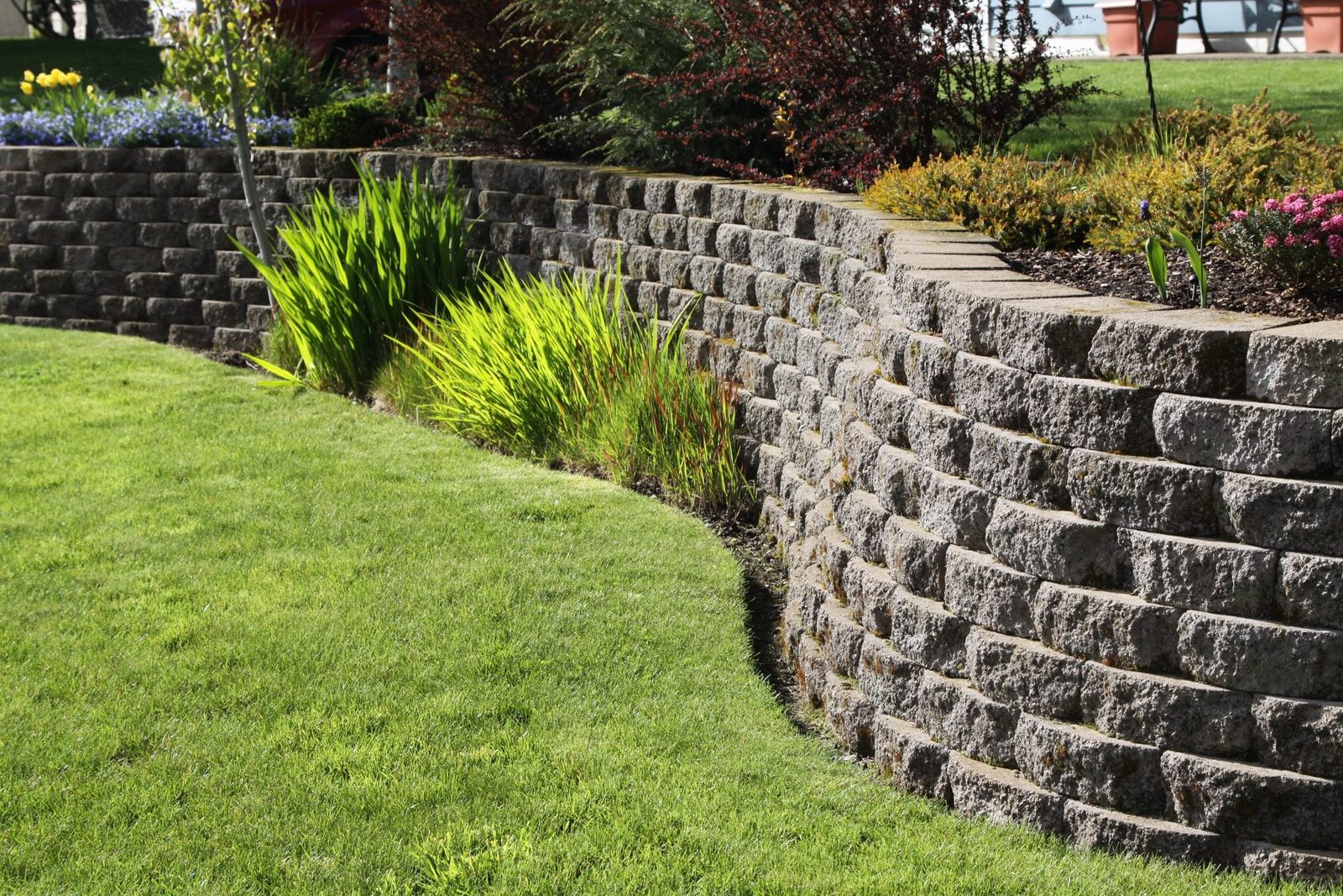 A curved stone retaining wall supports a lush green lawn, bordered by vibrant plants and flowers in a sunny garden setting.