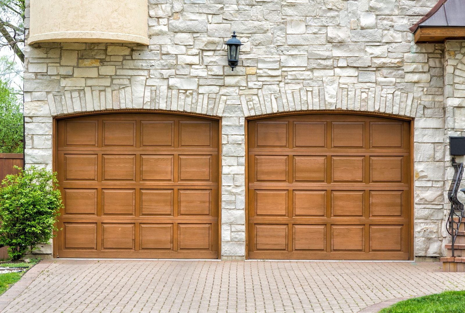 Two wooden garage doors are closed, standing against a stone wall, alongside a landscaped area with a small bush and a paver driveway.