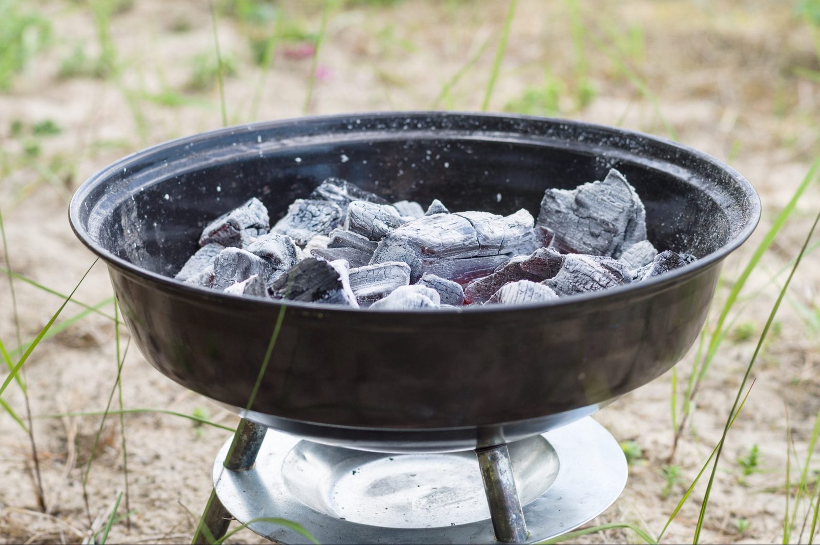 A charcoal grill contains glowing coals, preparing for cooking in an outdoor sandy area surrounded by sparse green grass.
