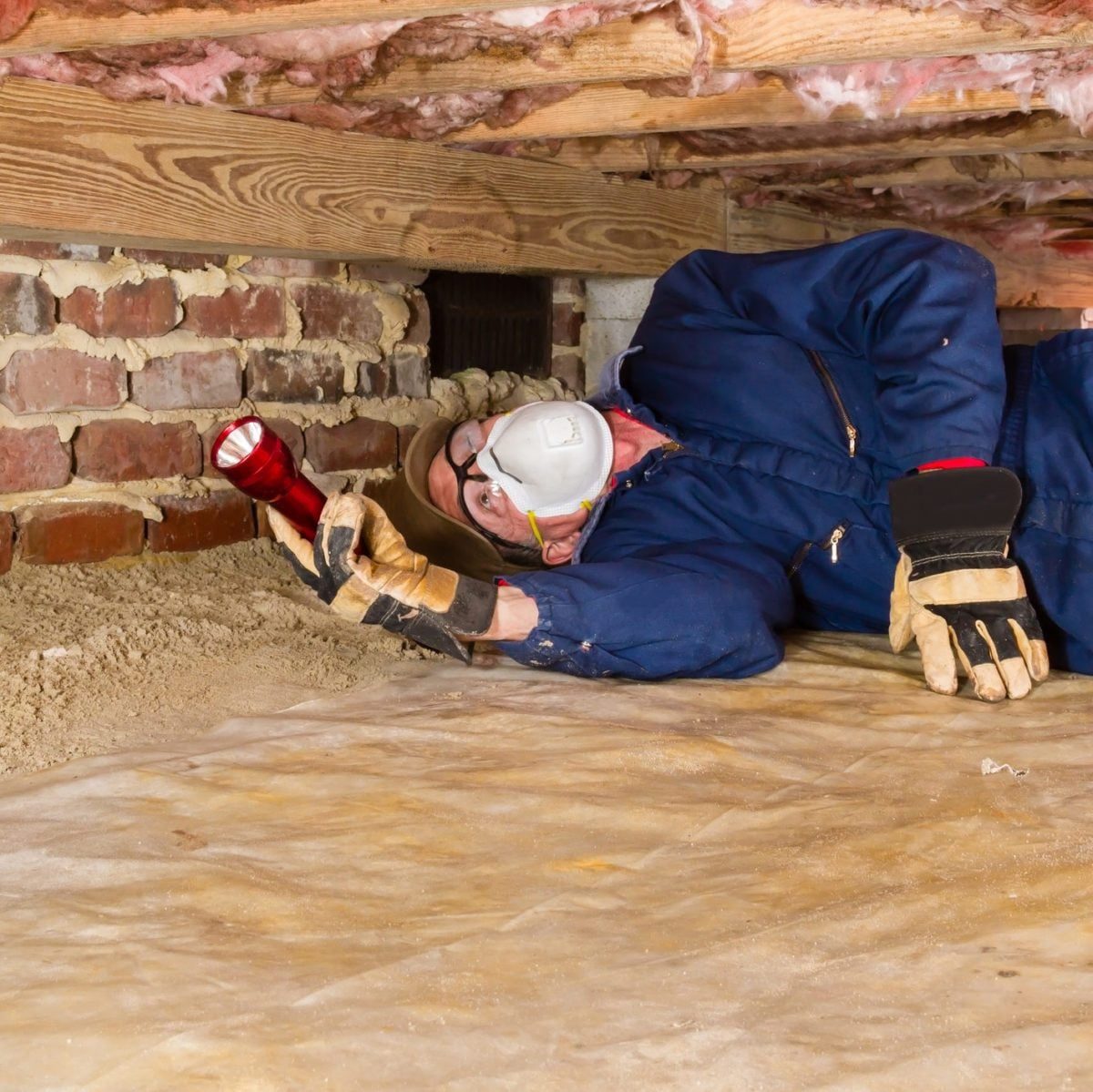A person in safety gear lies on a dusty floor, using a flashlight to examine a basement area between wooden beams and brick walls.