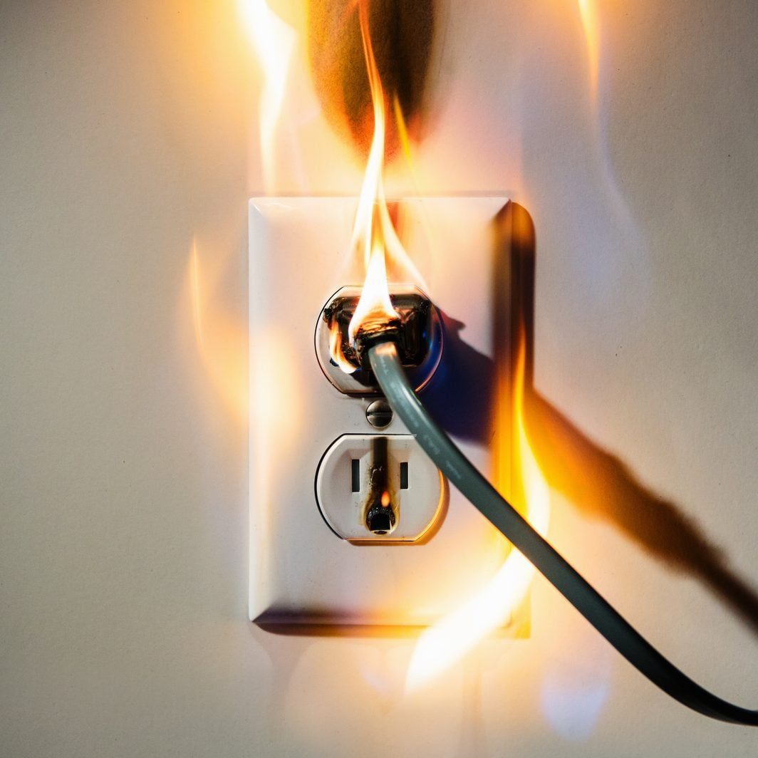An electrical outlet is on fire, with flames engulfing a plugged-in wire, set against a plain wall, indicating a potentially dangerous situa