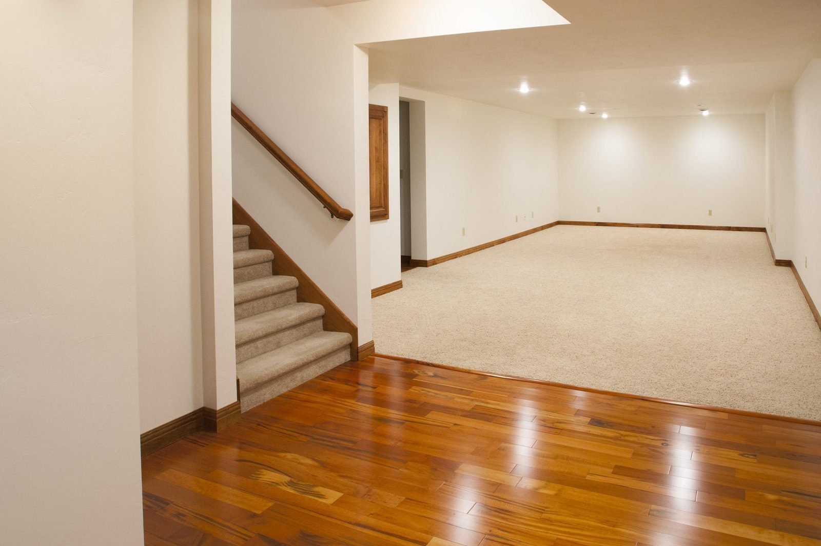 A staircase ascends from polished wooden flooring into a spacious, empty room with soft carpet and bright overhead lights in a neutral-color