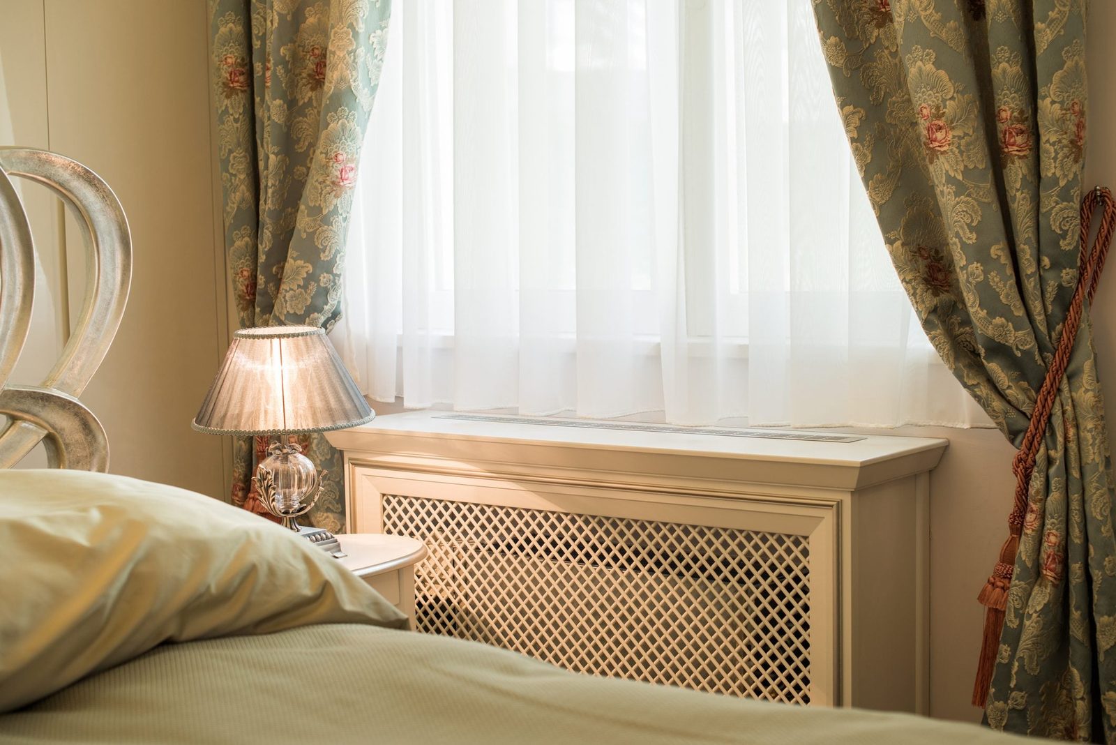 A bedside lamp illuminates a textured wall and elegant curtains, with soft bedding visible nearby, creating a cozy, serene bedroom atmospher
