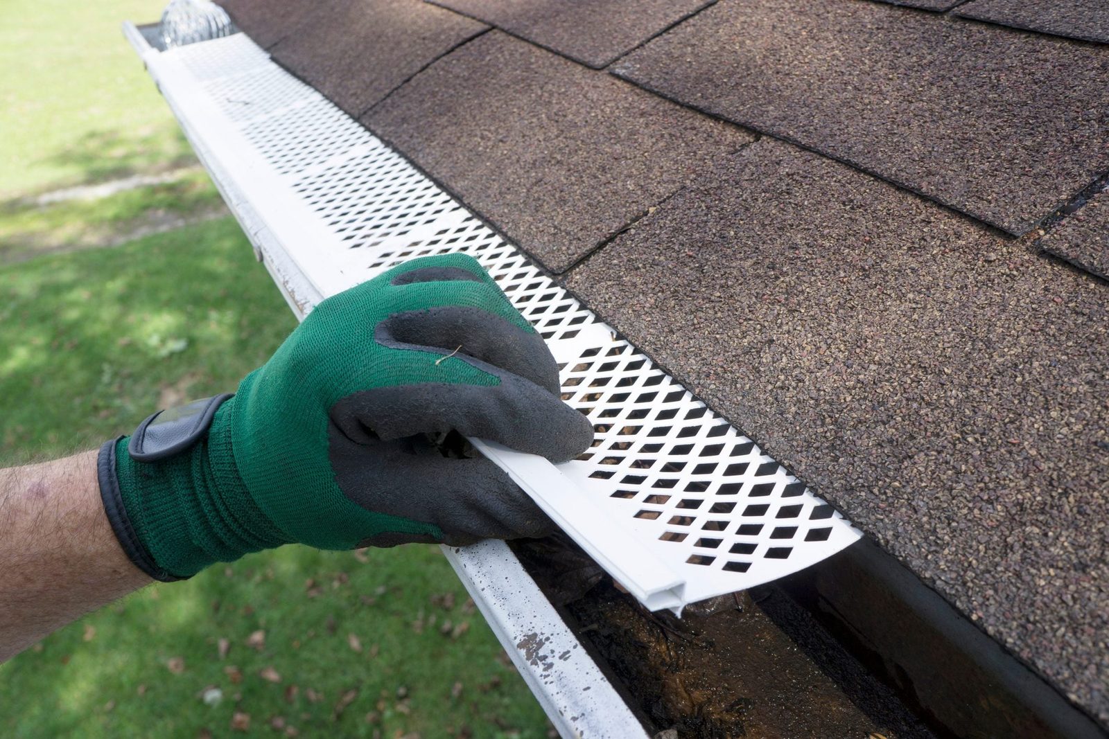 A gloved hand installs a white gutter guard on a roof, protecting the gutter from debris, with green grass visible in the background.
