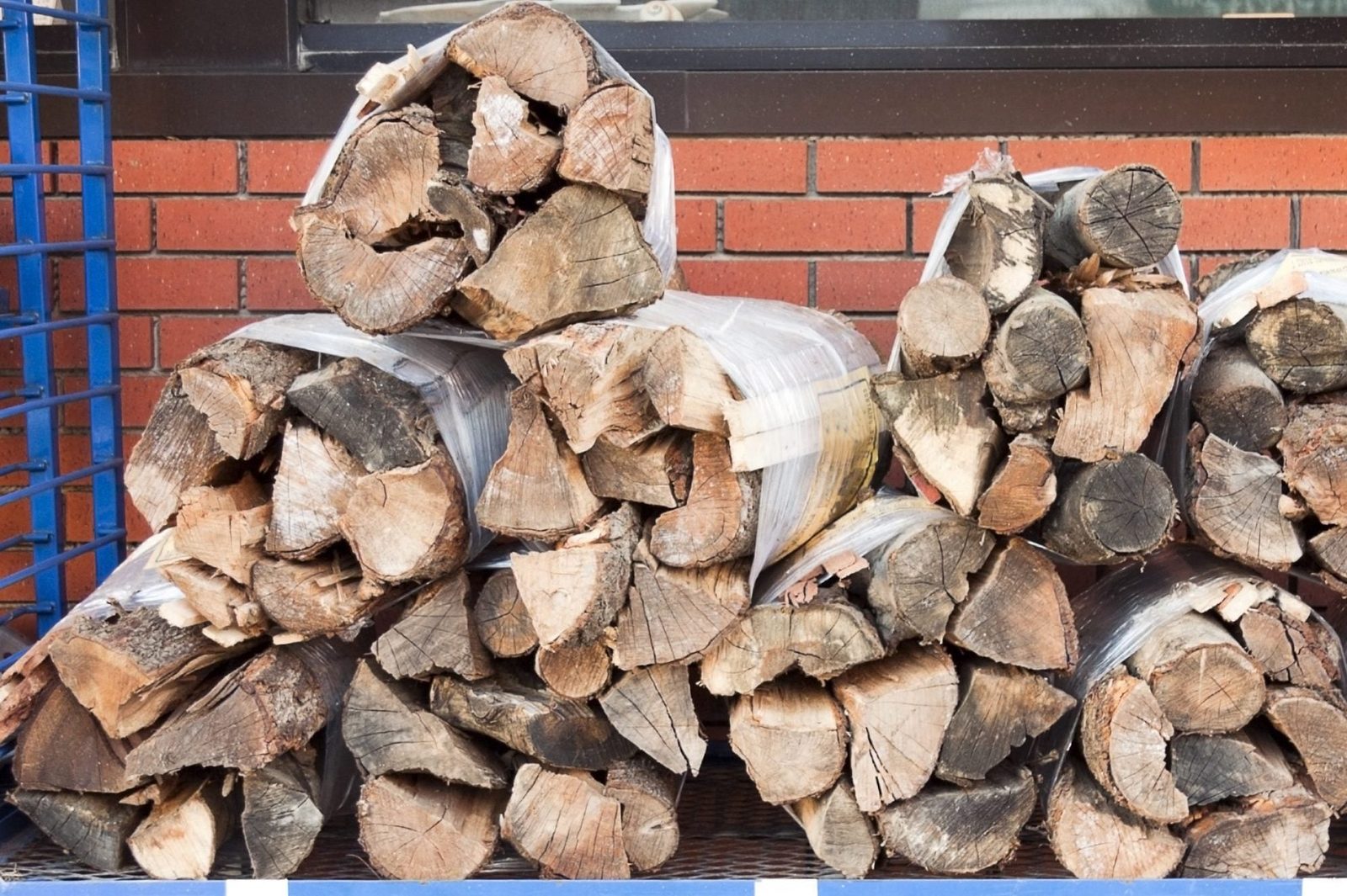 Stacked logs are wrapped and organized, resting against a brick wall in a structured outdoor display, likely for sale or storage.