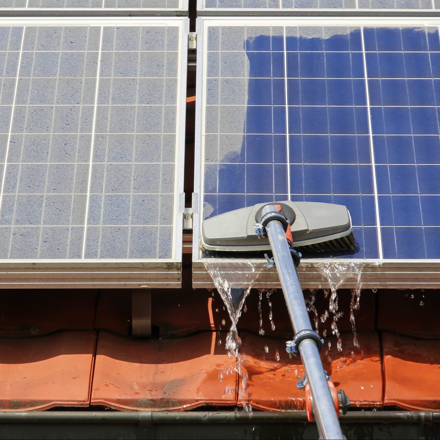A cleaning brush is being used to wash solar panels, with water flowing off the panels onto a red-tiled roof below.