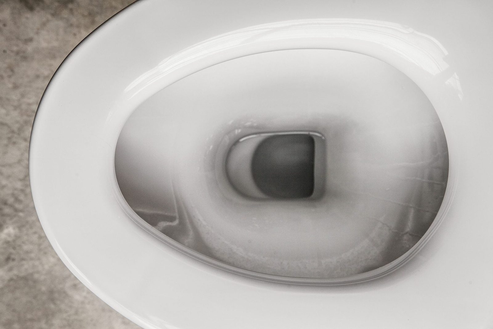 A white toilet bowl is positioned centrally, filled with clear water, against a textured gray concrete surface, reflecting overhead light.