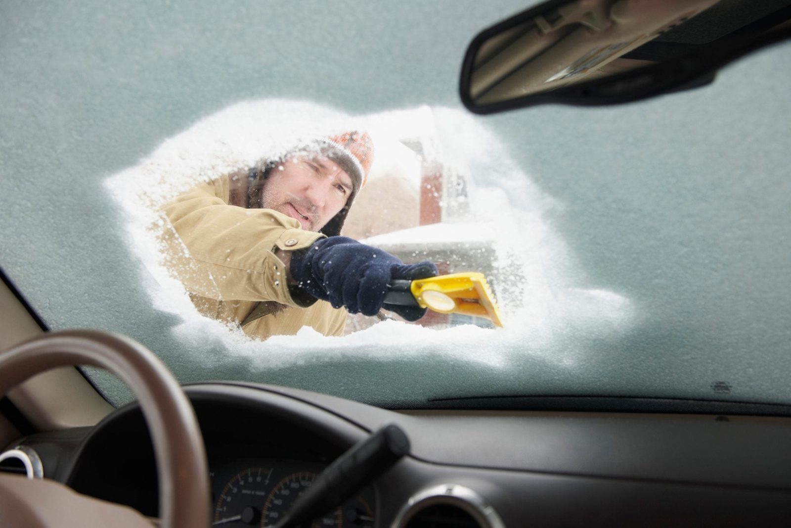 A person is scraping snow off a car's windshield with a yellow tool while inside the vehicle, surrounded by a wintry environment.
