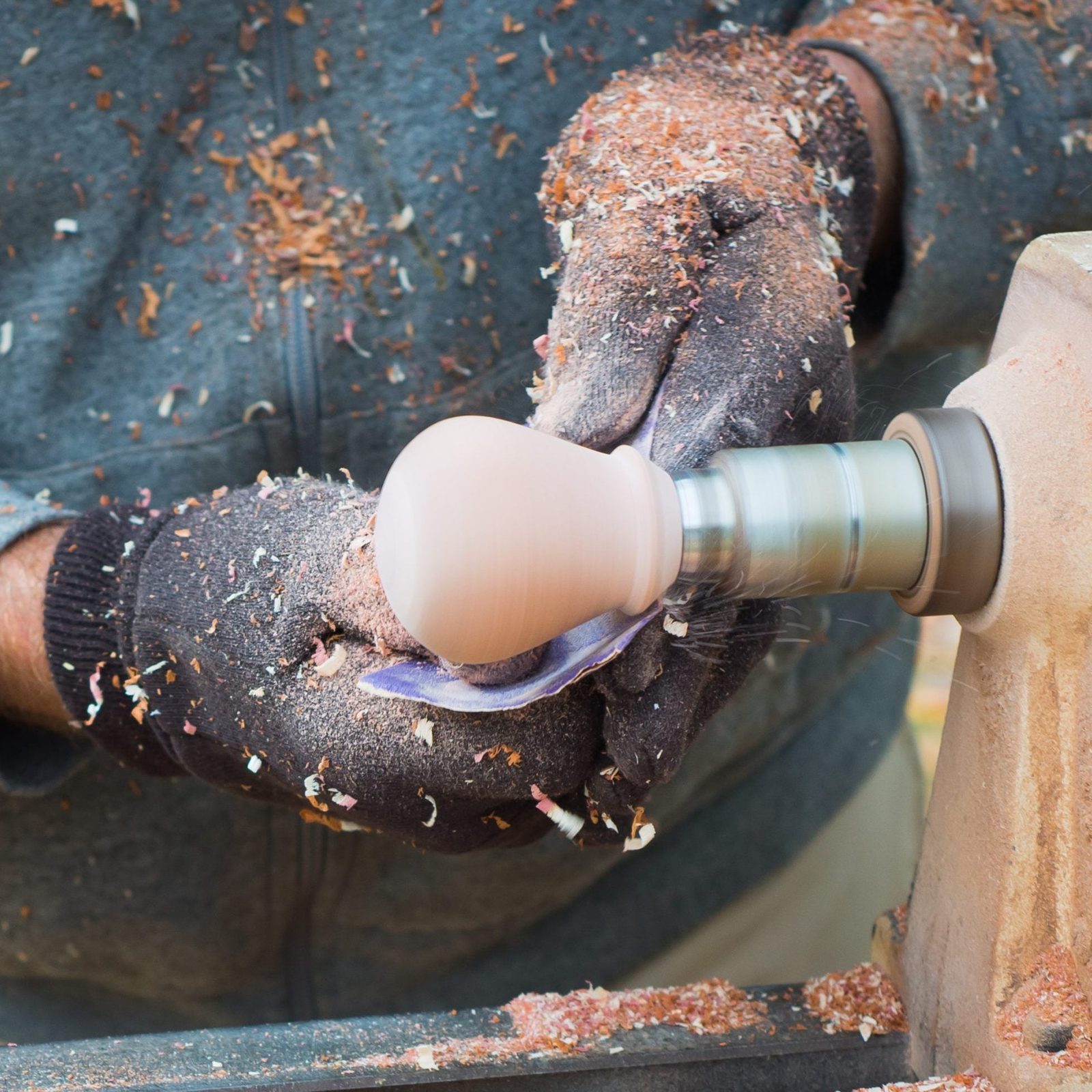 A craftsman, wearing gloves, shapes a wooden piece on a lathe, surrounded by wood shavings and particles in a workshop setting.
