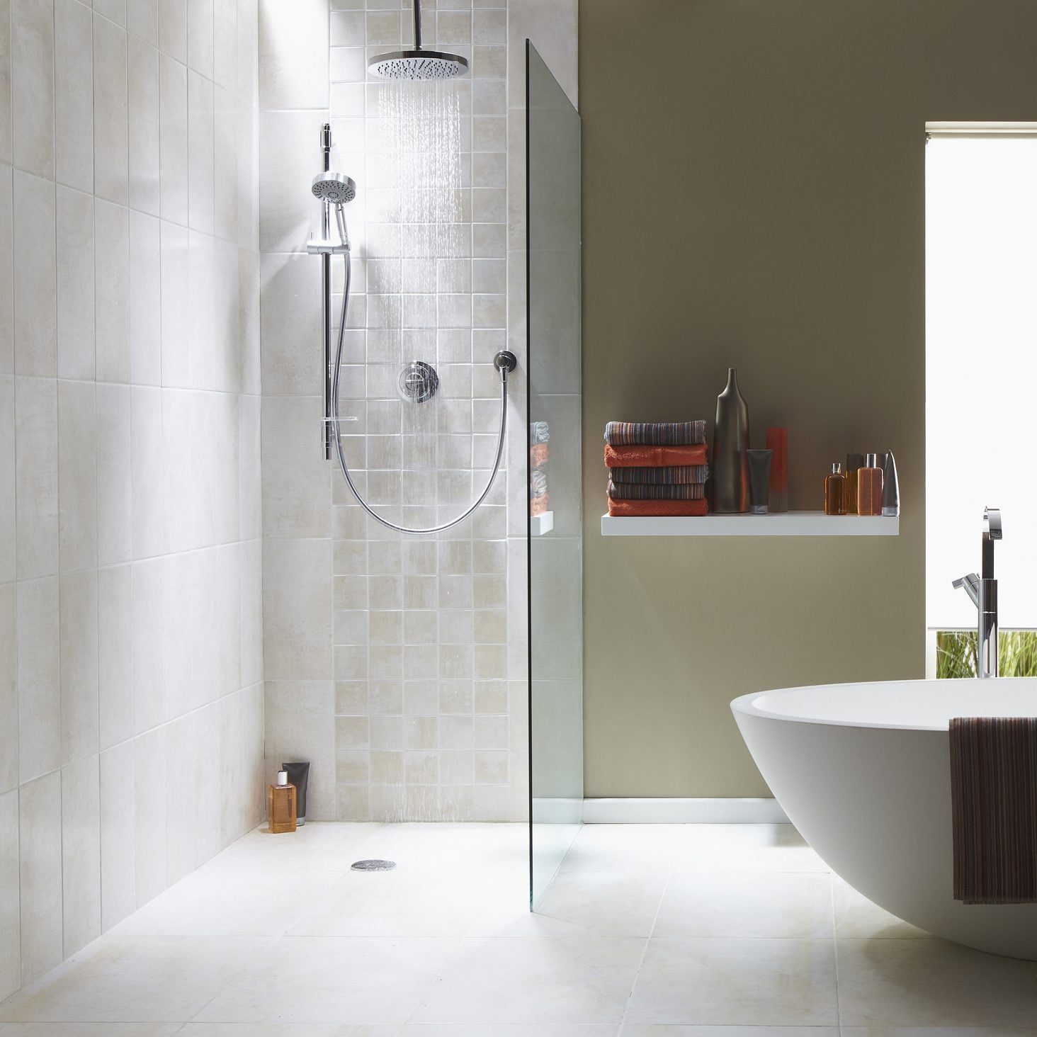 A showerhead releases water in a bright bathroom, with a glass partition and a white bathtub nearby, surrounded by neatly stacked towels and