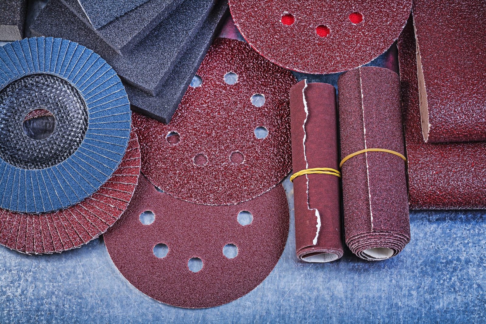 Various sanding materials, including discs and sheets, are stacked and arranged on a textured blue surface, demonstrating their diverse shap