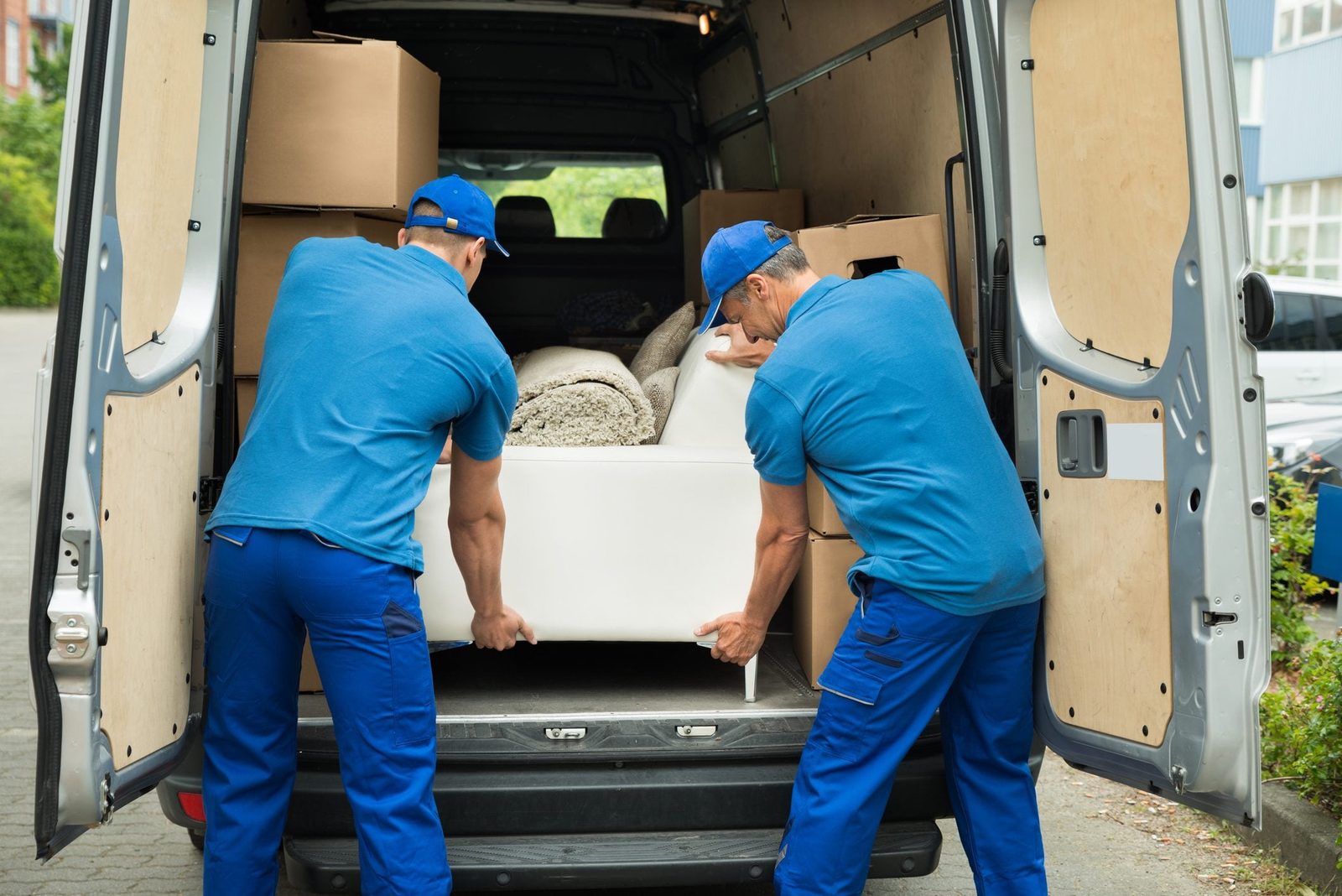 Two workers lift a large white object from a van, surrounded by boxes and rugs, in a parking area with greenery in the background.