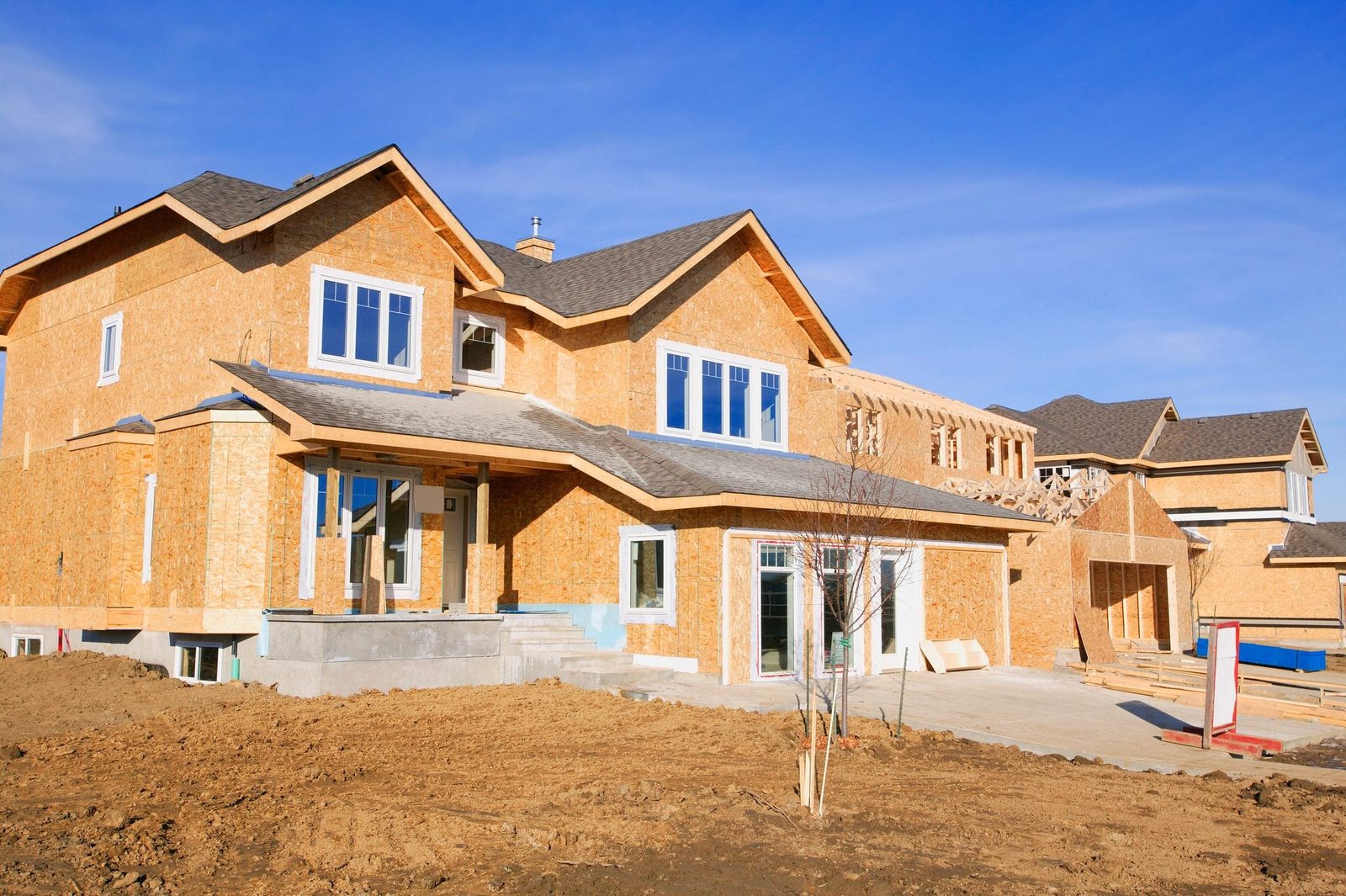 A partially constructed house features wooden framing, large windows, and an unfinished exterior, situated on bare soil under a clear blue s
