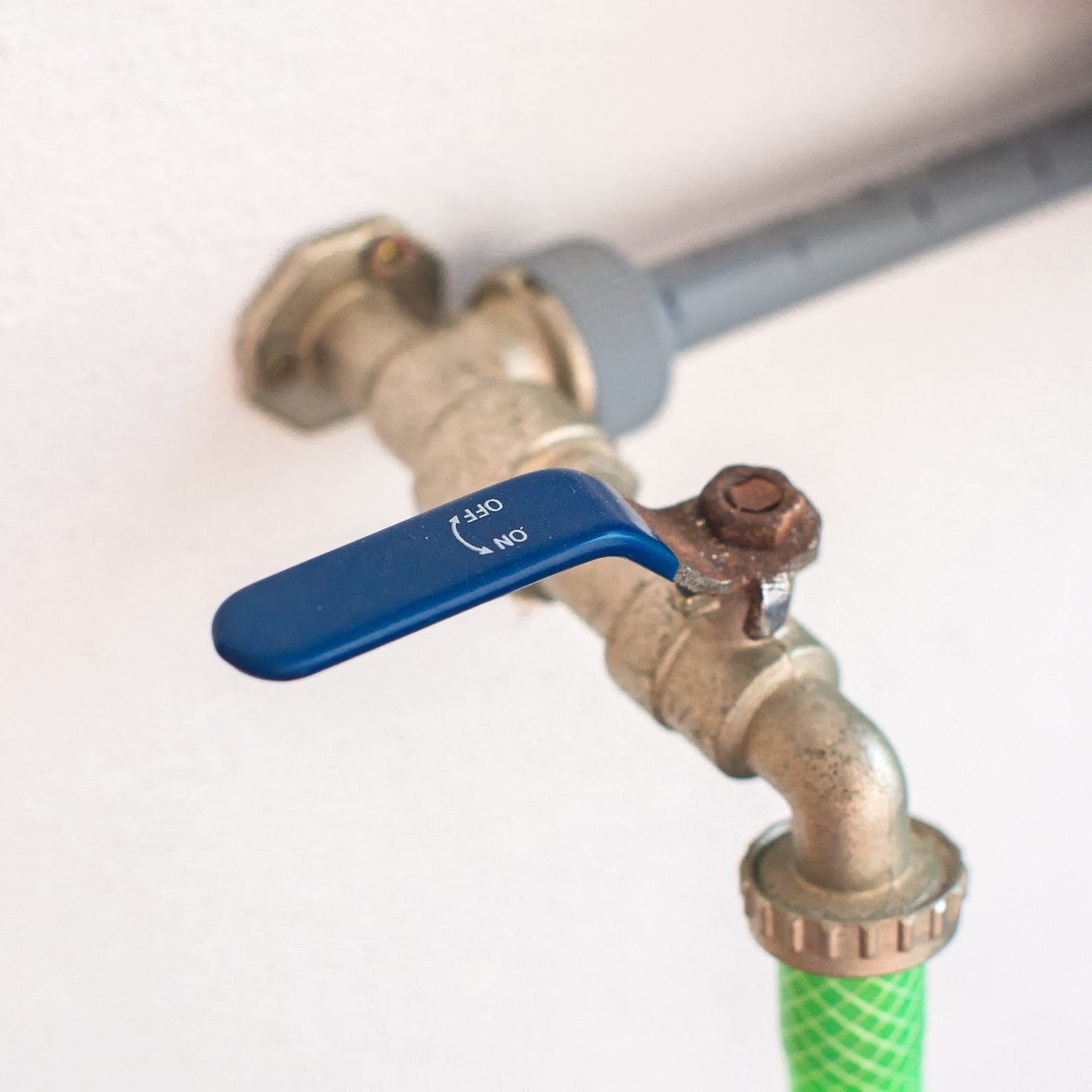 A blue valve is turned to control water flow, connected to a metallic pipe with a green hose attached, set against a white wall.