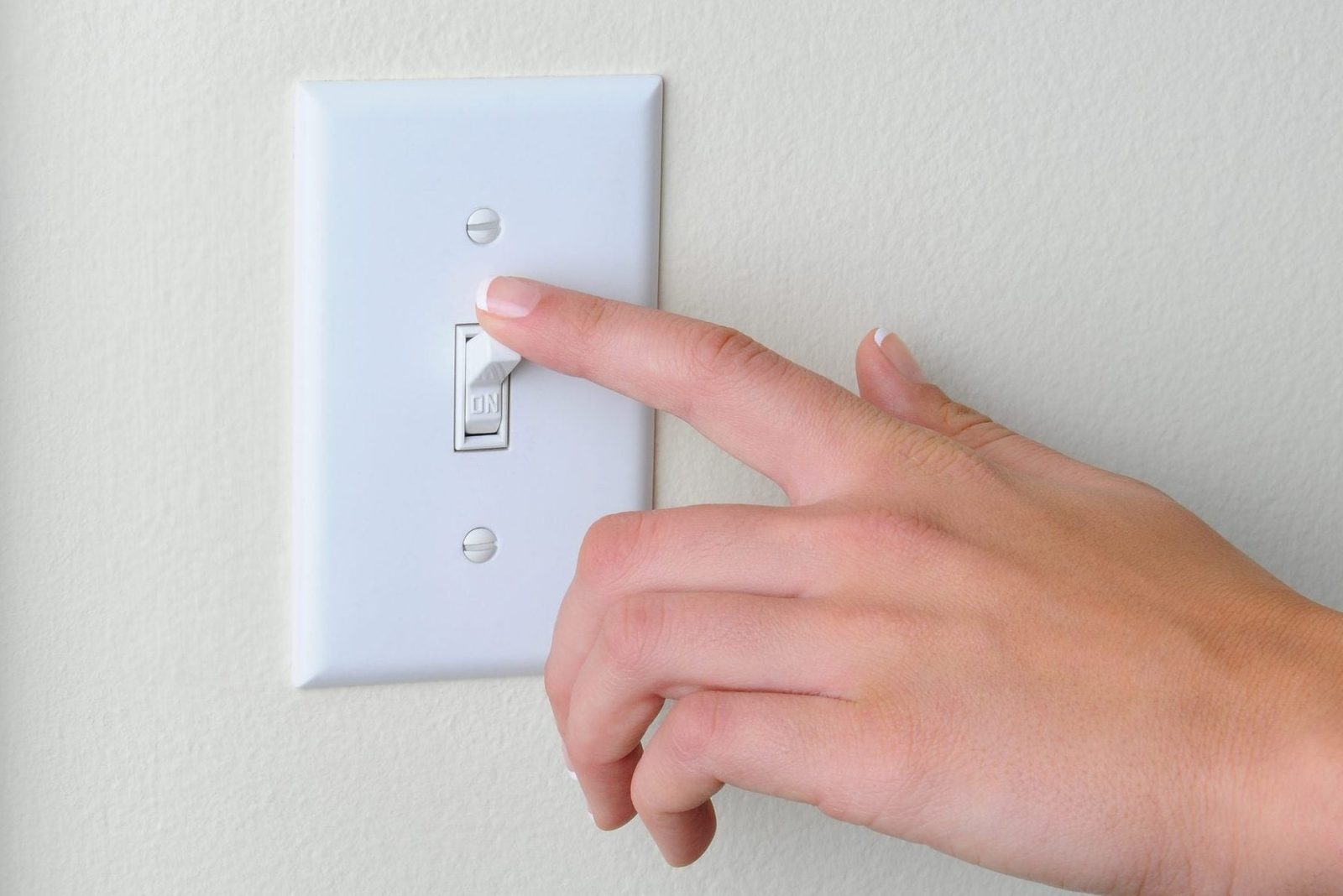 A hand is pressing a white light switch labeled "ON" on a plain wall, indicating the action of turning on a light.