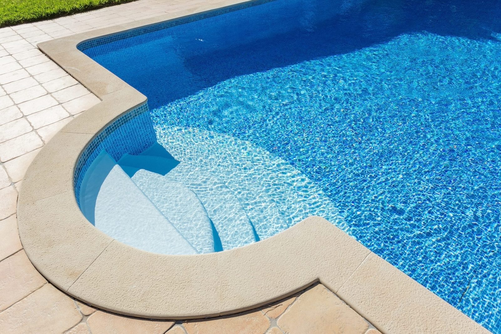 A rounded swimming pool glistens under sunlight, featuring blue water and white steps leading into it, surrounded by a tiled deck and green
