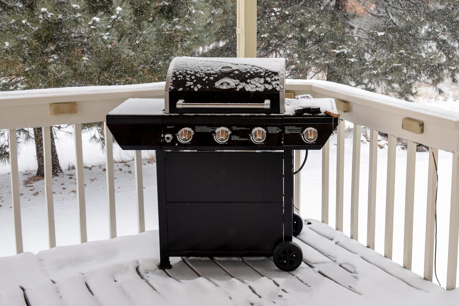 A black gas grill covered in snow sits on a wooden deck, surrounded by snow-covered ground and trees, while snowflakes fall gently.