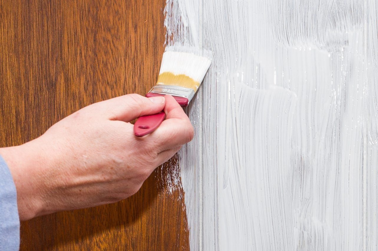 A hand paints a section of wood with a brush, applying white paint. The contrasting wood grain is visible beside the painted area.