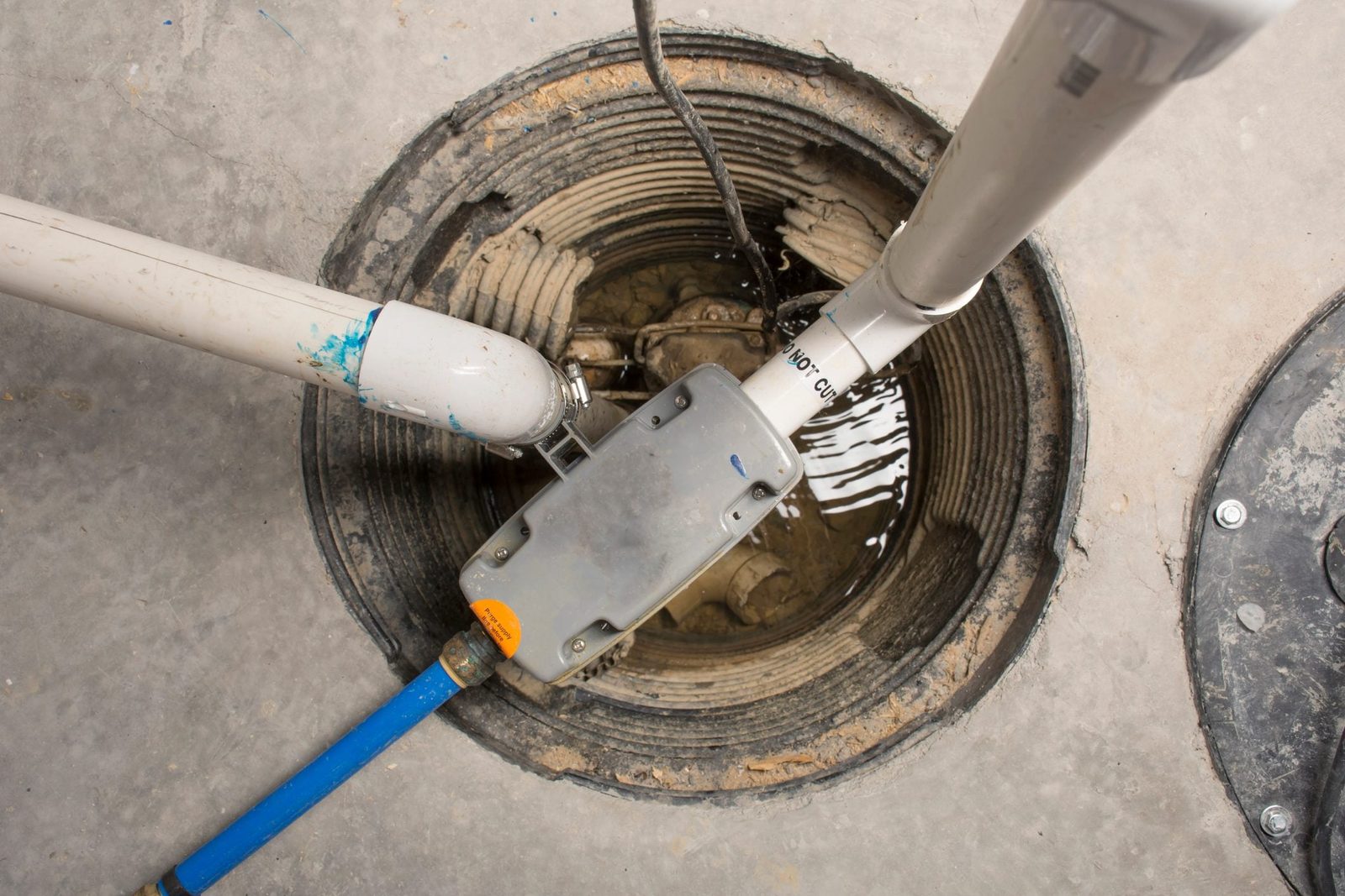 A sump pump is positioned in a circular basin, surrounded by concrete, with pipes directing water in and out, sitting above murky water.