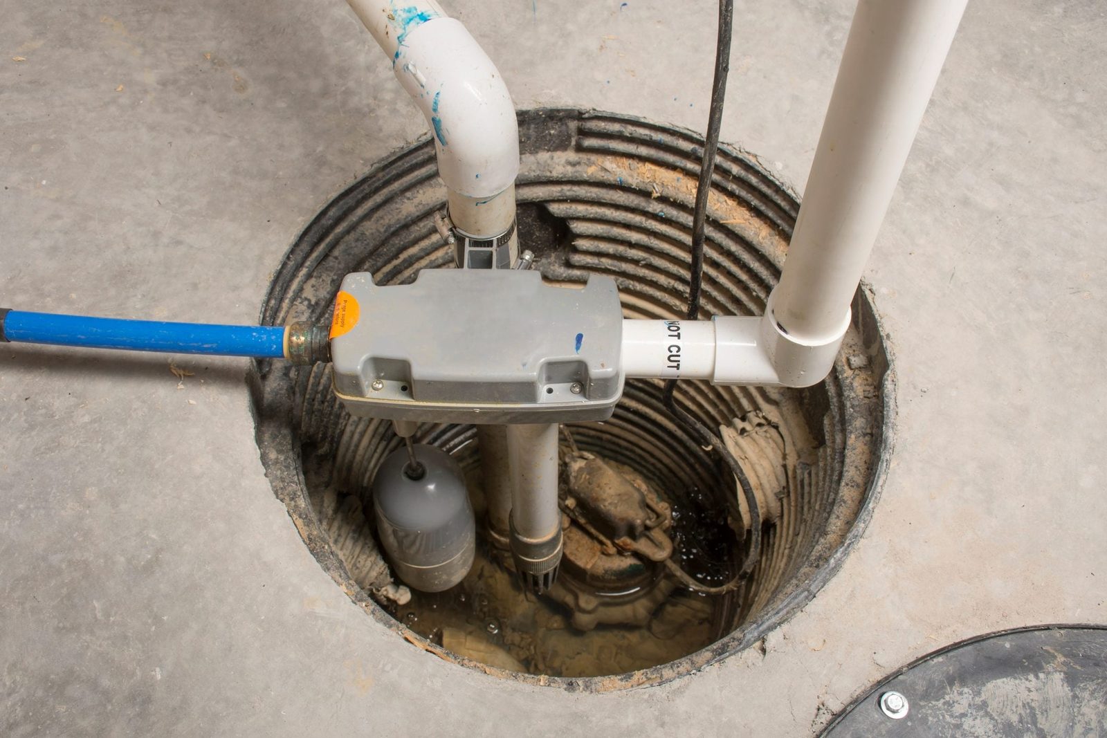A sump pump is positioned in a cylindrical pit, pumping water from a wet environment into an external drainage system. Visible pipes connect above the pump.