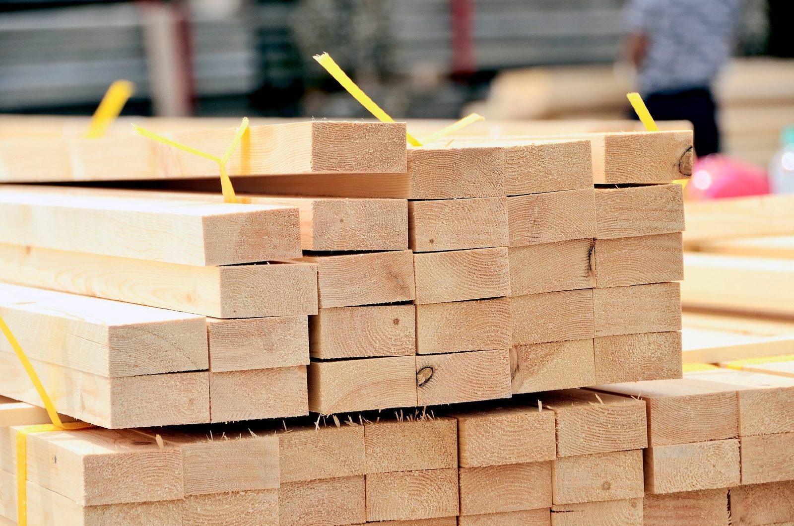 Stacked wooden planks are bound with yellow straps, resting on a construction site, surrounded by other materials and equipment.