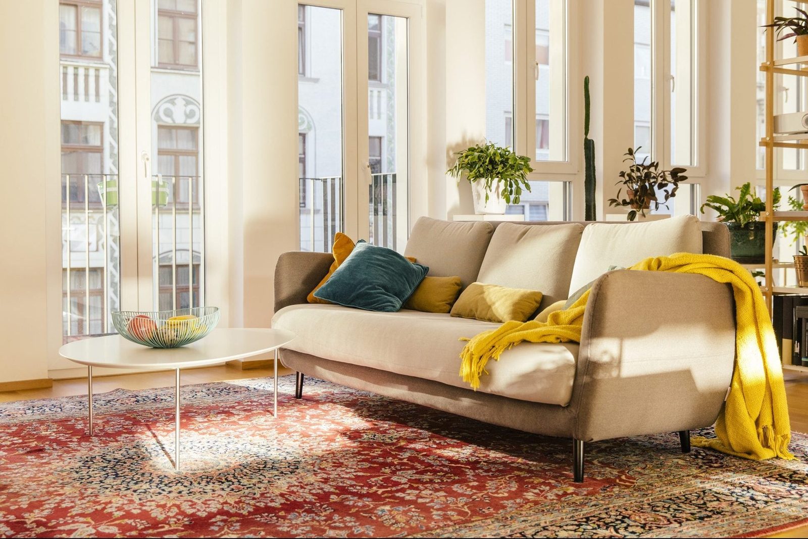 A sofa adorned with colorful pillows rests on a patterned rug, beside a coffee table with a bowl of fruit, illuminated by sunlight through l