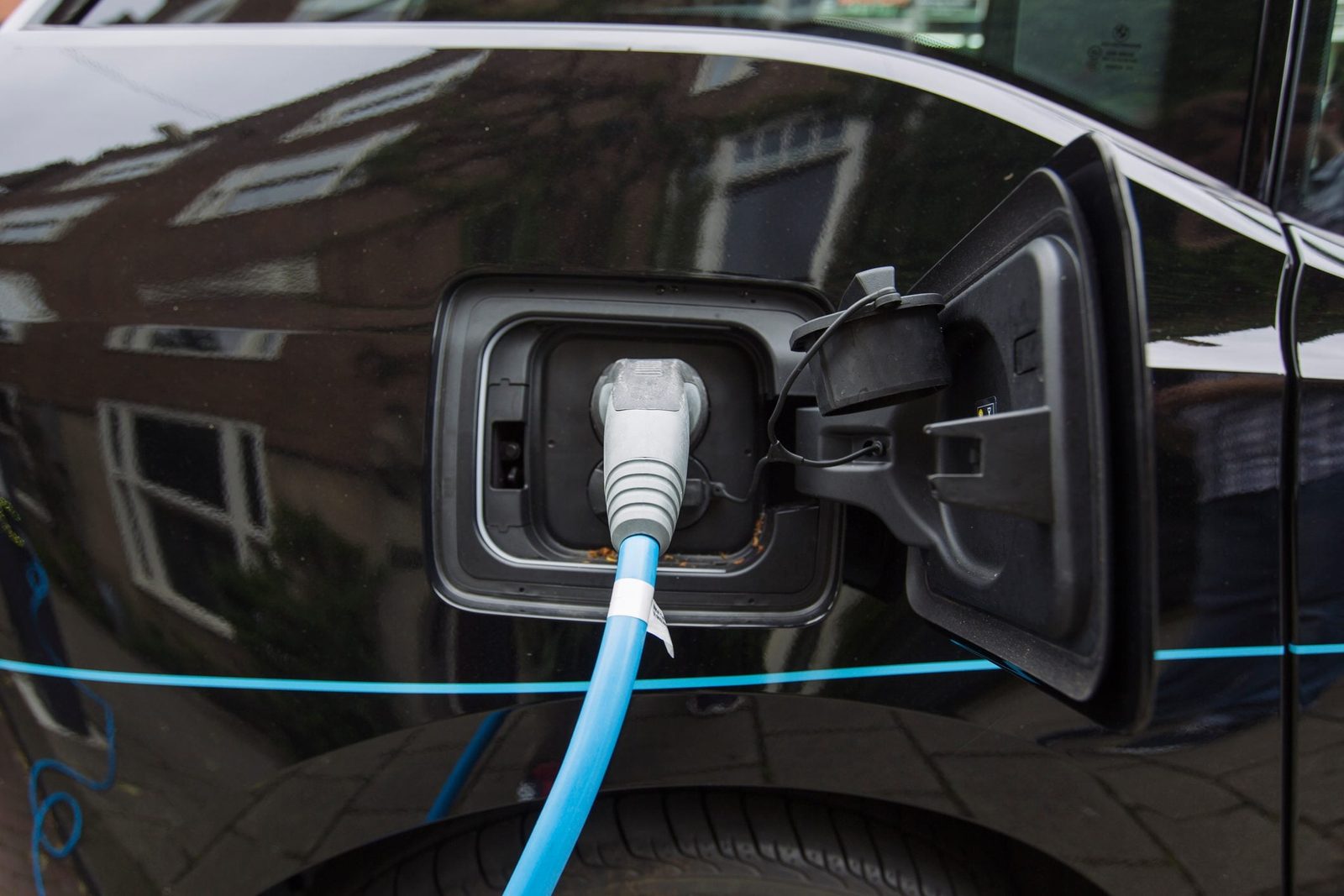 An electric car's charging port is connected to a blue charging cable, situated on a black vehicle parked in an urban environment with build