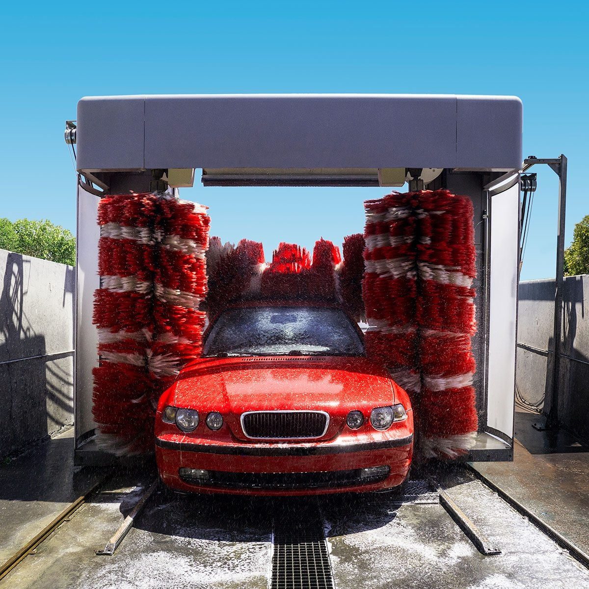 A red car is being cleaned in a car wash, surrounded by rotating brushes spraying water, under a clear blue sky with greenery in the backgro