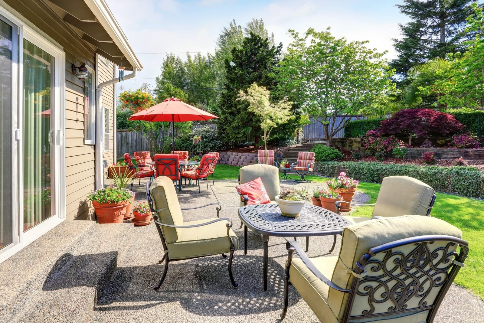 A round table with chairs sits on a patio. Potted plants surround it, while bright umbrellas shade distant seating in a vibrant garden.