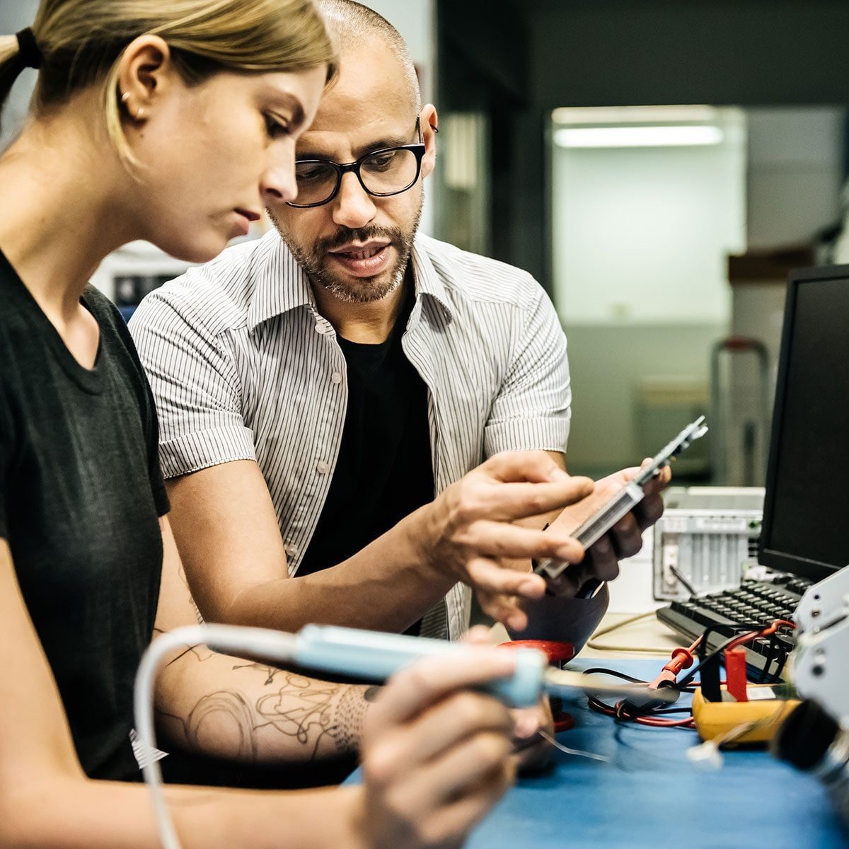 Two people, a man and a woman, are focused on a device being repaired at a cluttered workspace, sharing technical insights while surrounded