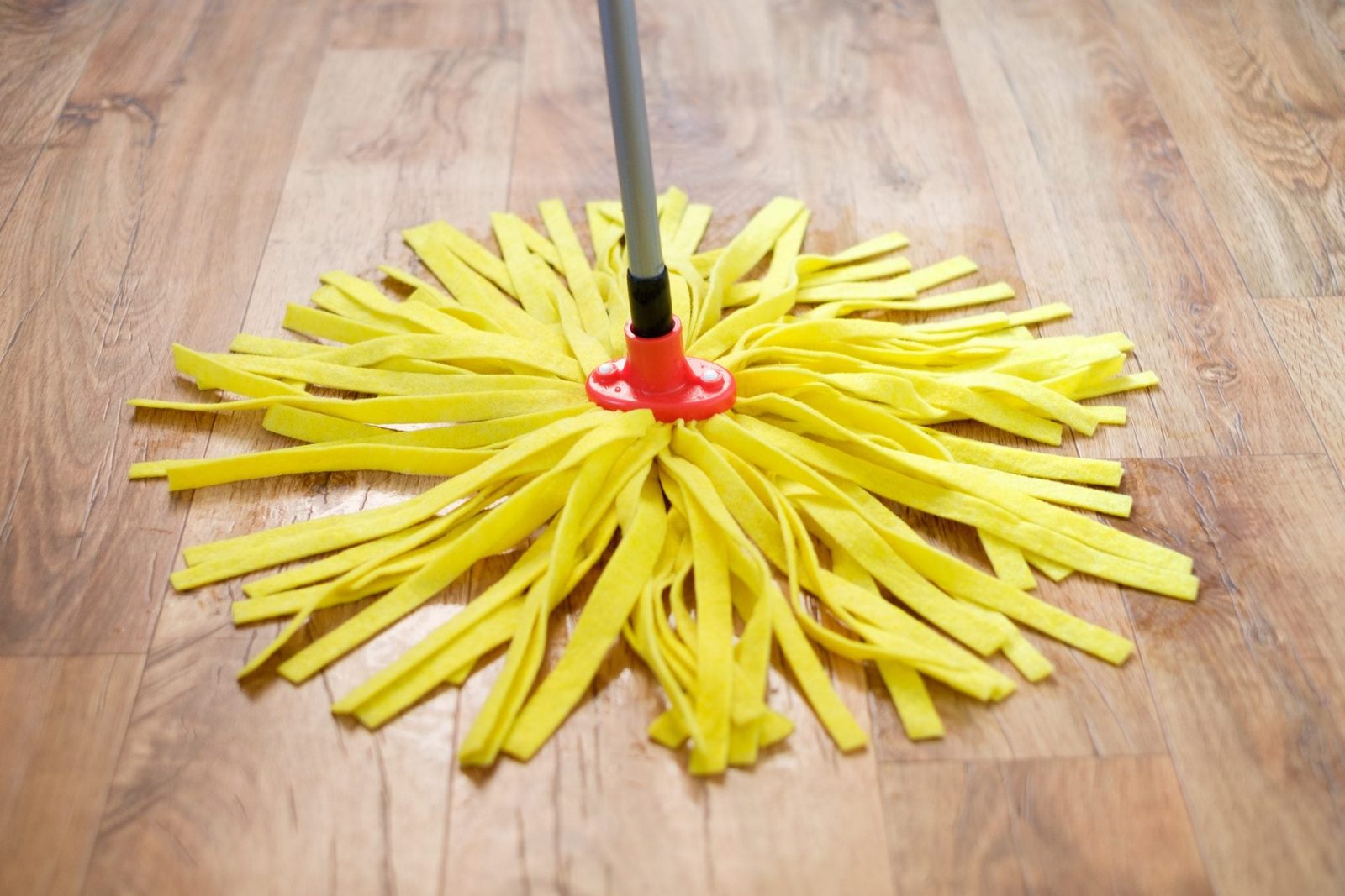 A yellow mop head sits on a wooden floor, with long strands radiating outwards, suggesting readiness for cleaning a surface.