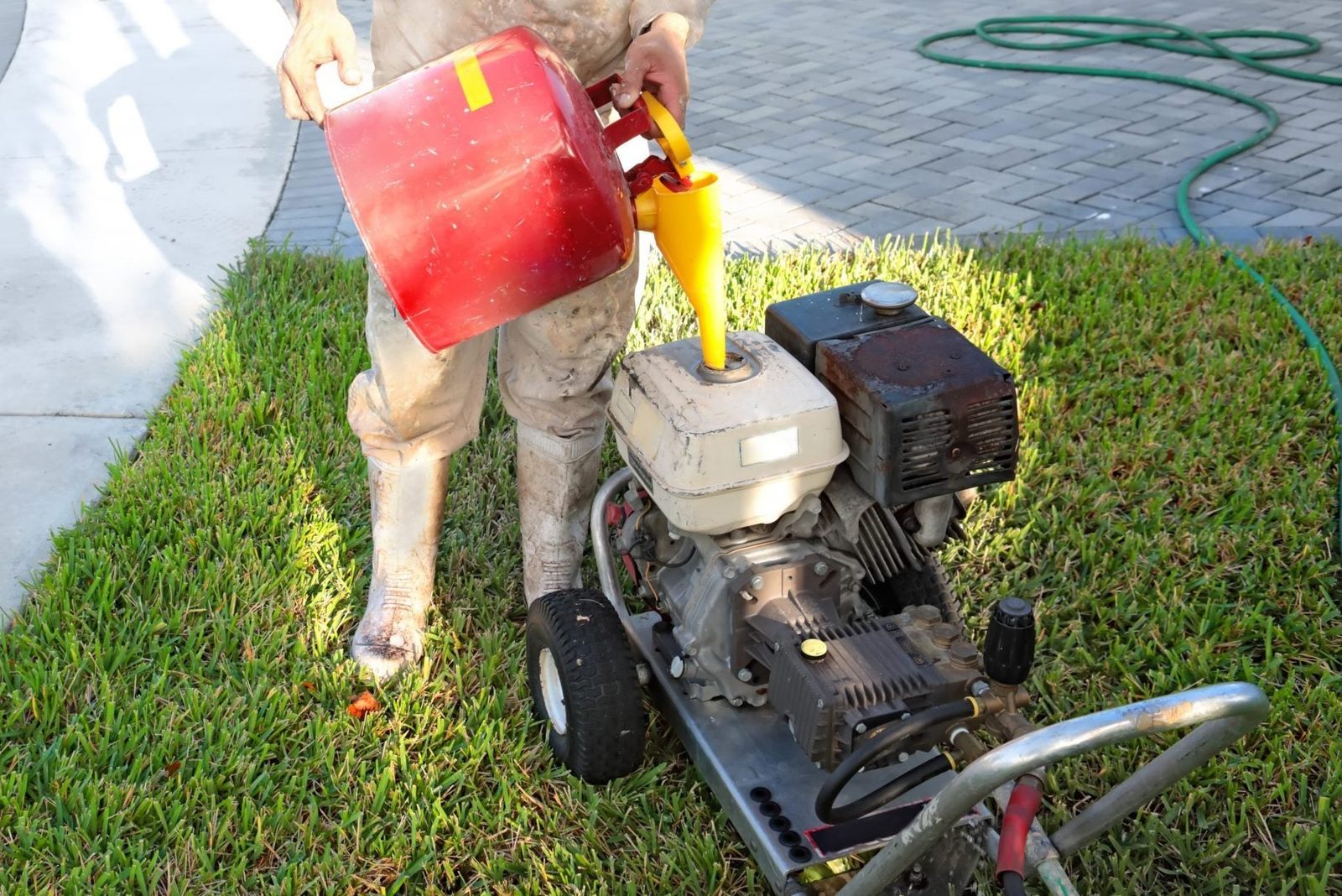 A person pours fuel from a red can into a gas-powered machine on green grass, near a paved pathway and a garden hose.