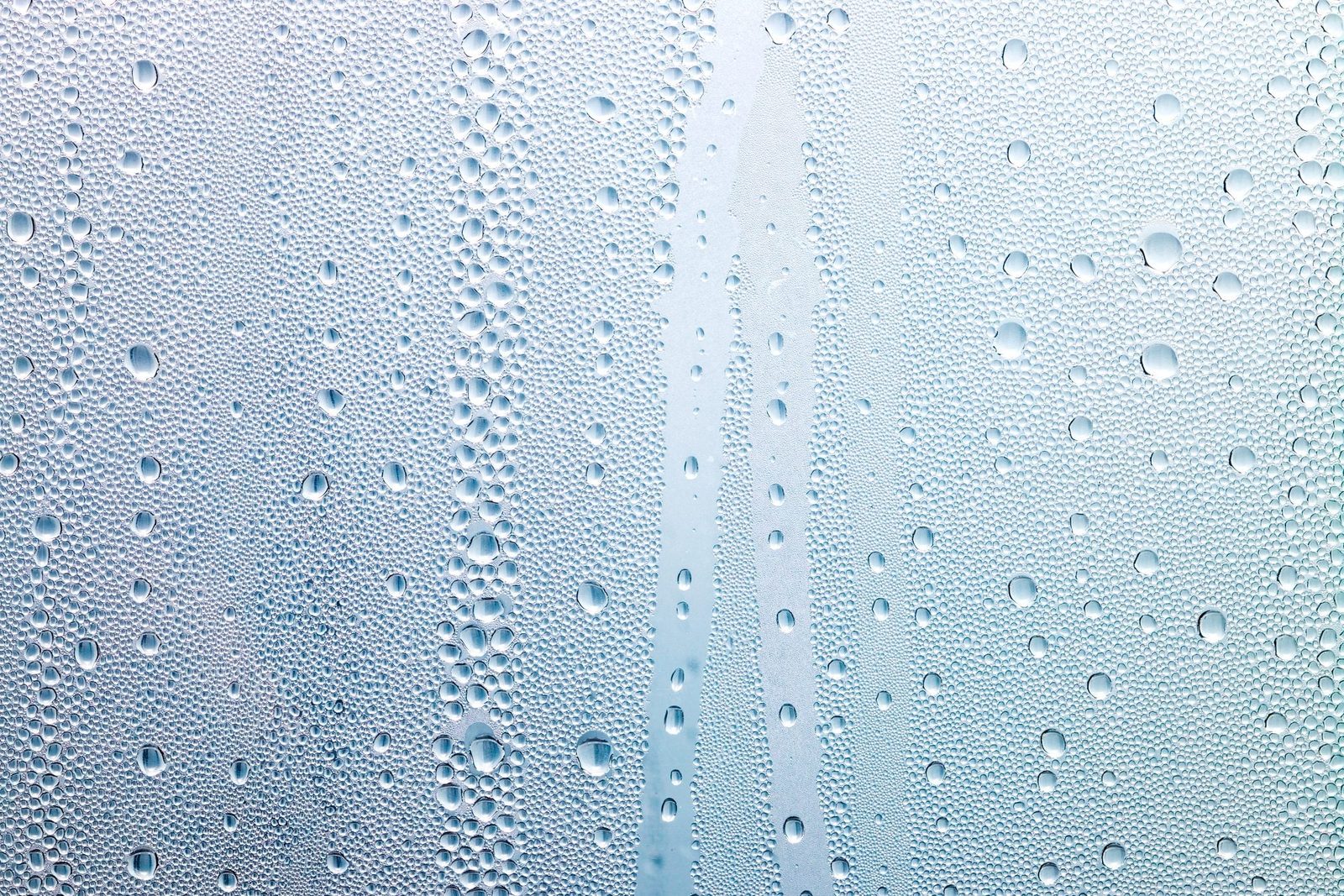 Water droplets cling to a smooth surface, glistening as they slowly slide down, with a light blue background enhancing the wet appearance.