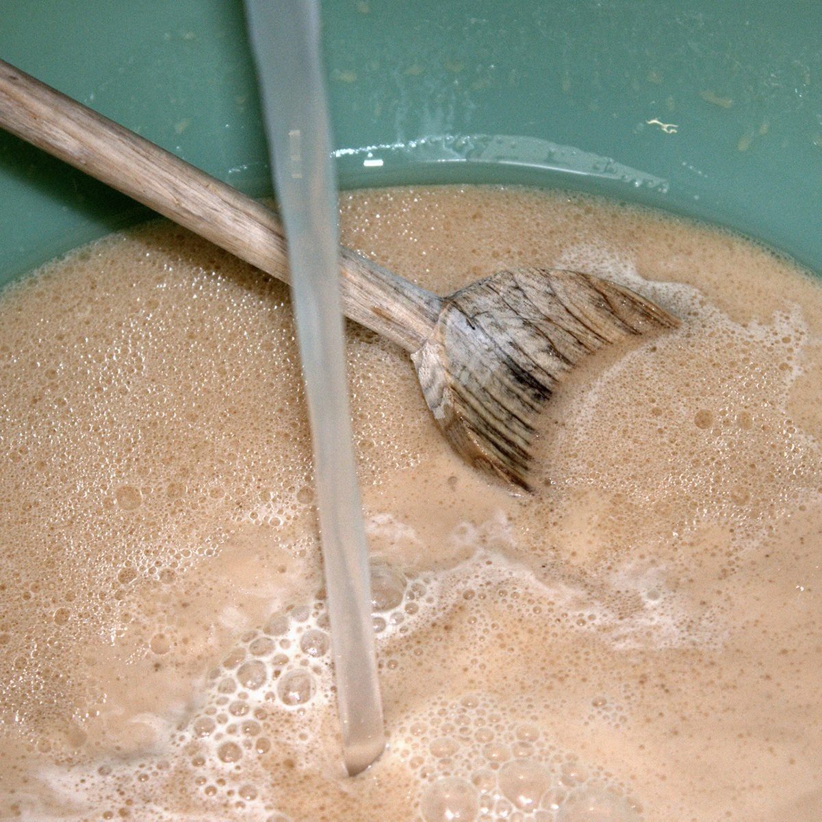 A wooden spoon stirs a foamy, beige mixture in a light blue bowl, while a clear liquid flows into the bowl, enhancing the froth.