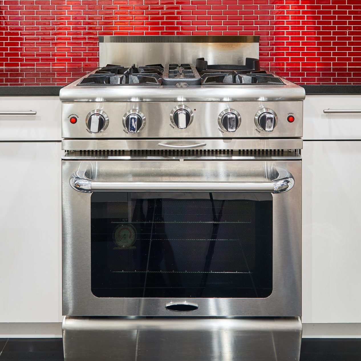 A stainless steel gas range with four burners is positioned in a modern kitchen, set against a vibrant red tiled backsplash.