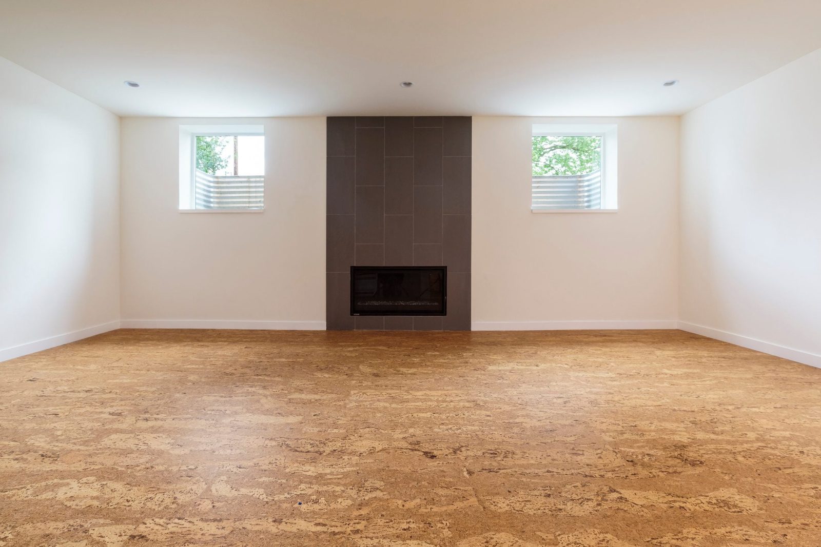 A fireplace is centered on a cork-floored room, illuminated by light from two windows on opposite walls, surrounded by plain white walls.