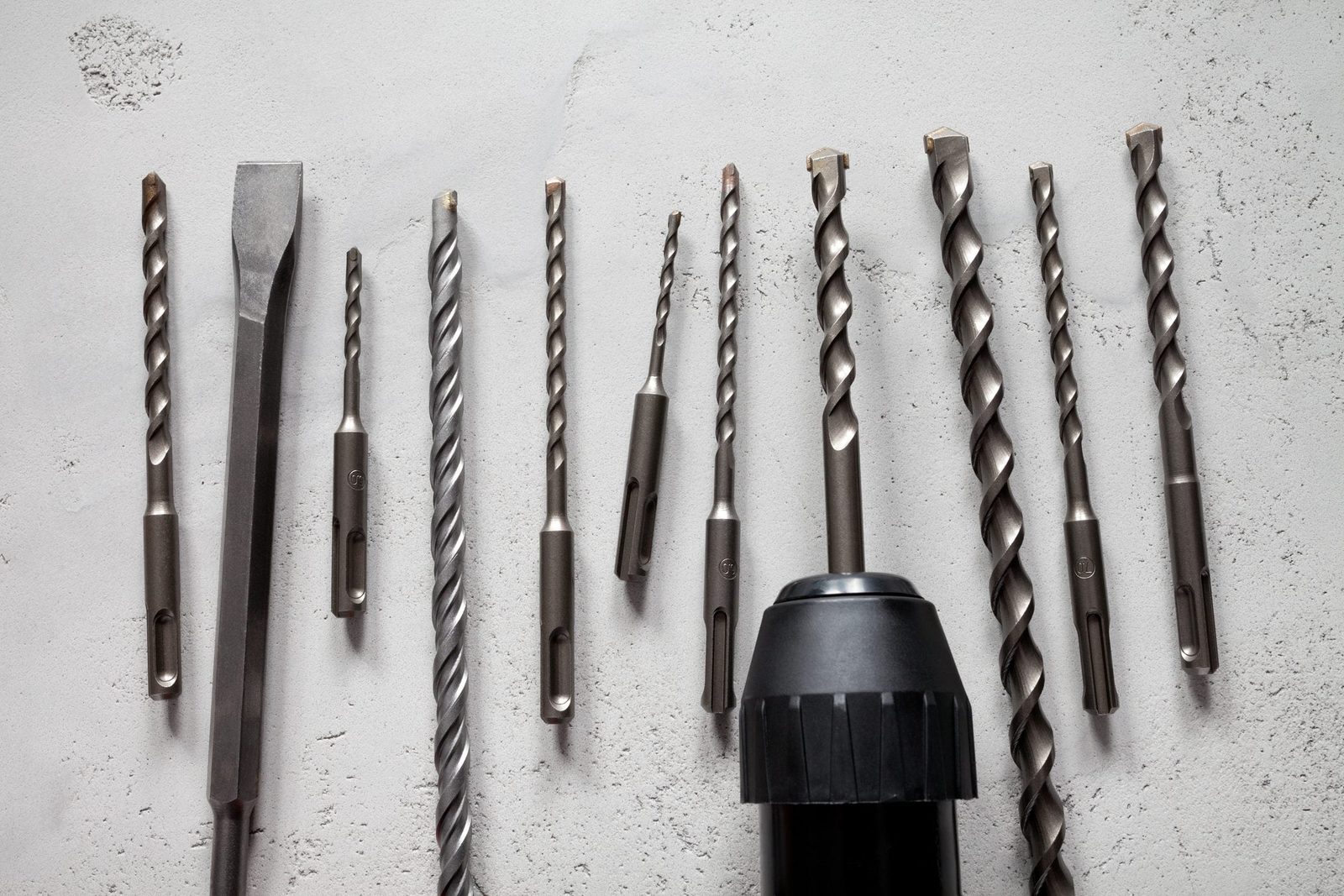 A collection of various drill bits and a drill are arranged on a light gray surface, showcasing their metallic texture and differing lengths