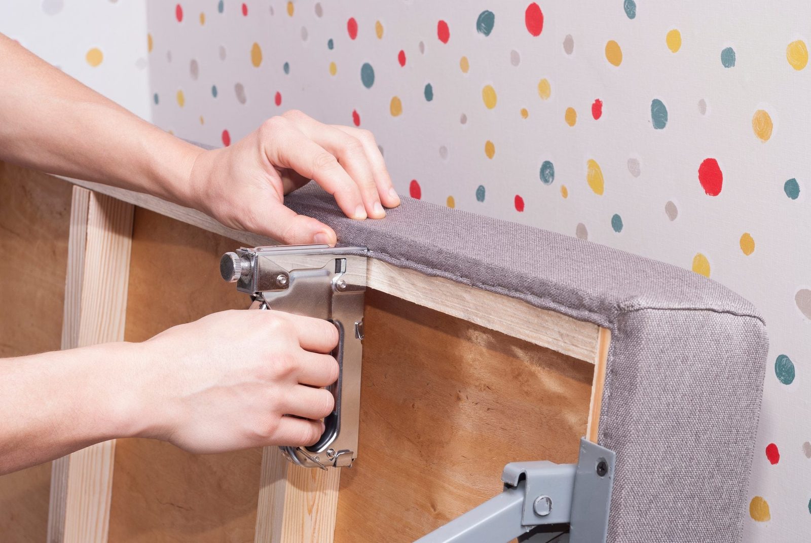 A person staples gray fabric onto a wooden frame, surrounded by a colorful, polka-dotted wall.