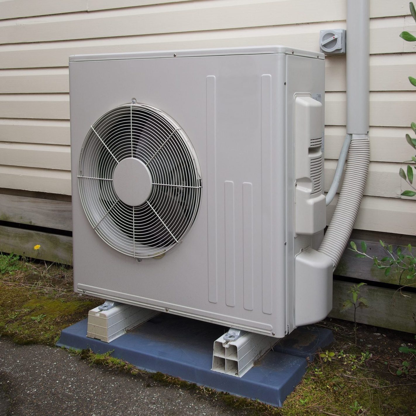 A gray outdoor air conditioning unit stands on a platform beside a wall, with a fan mounted on its side, in a garden setting.