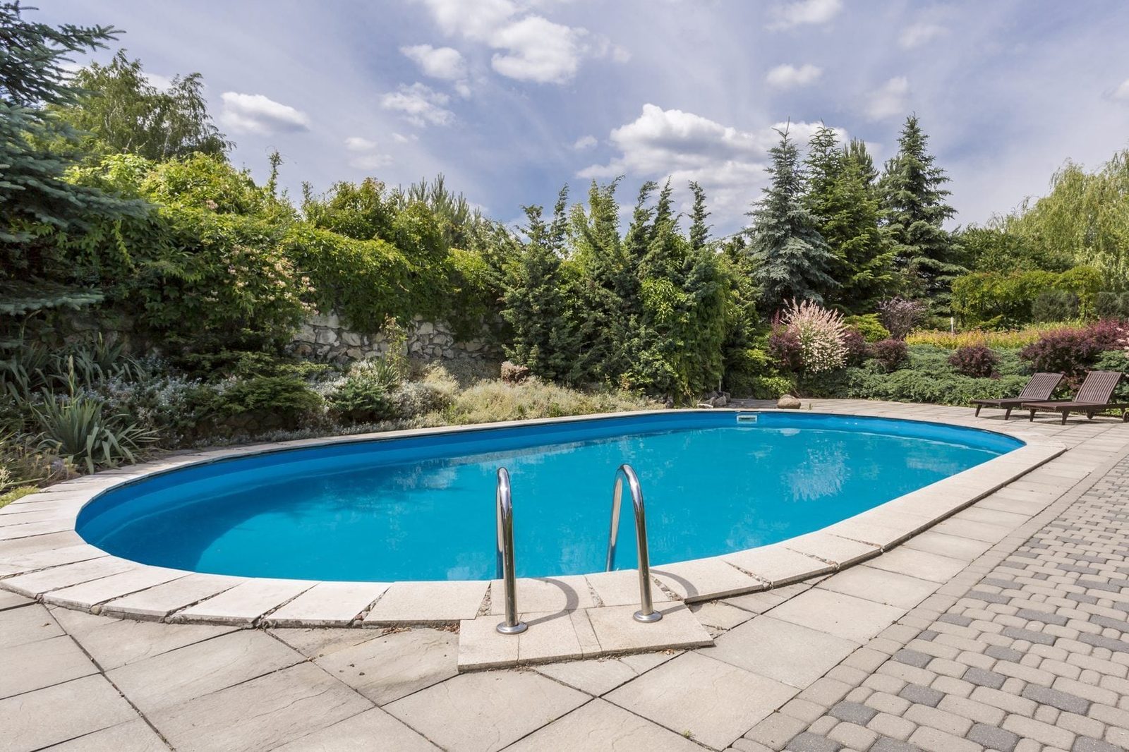 An oval swimming pool with metallic ladders sits on tiled flooring, surrounded by lush greenery and flowering plants under a partly cloudy s